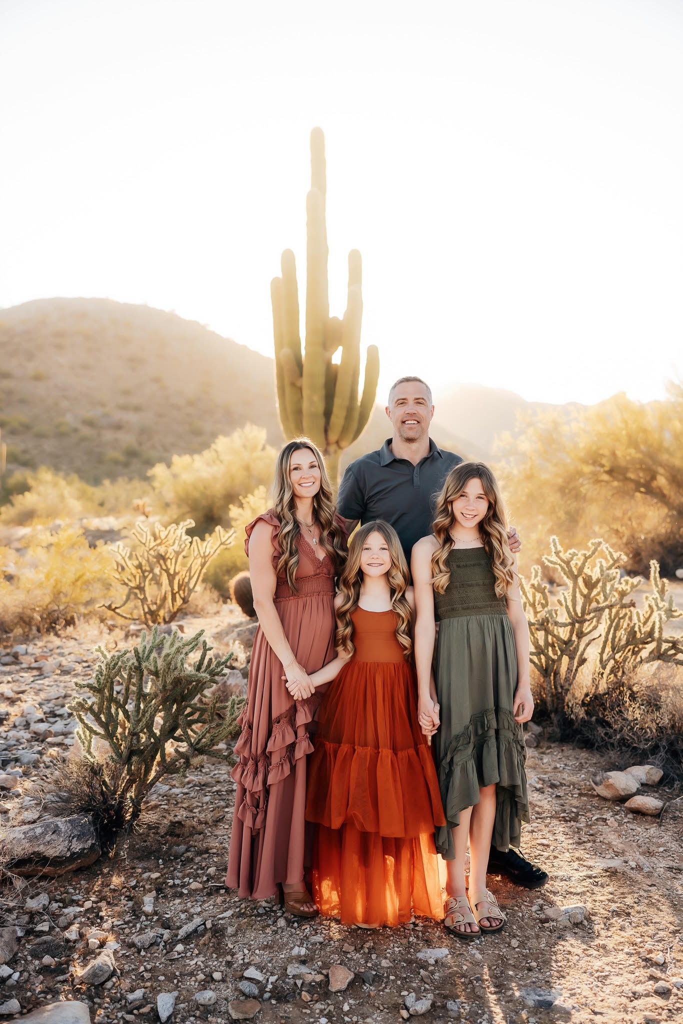 A family of five standing outdoors in a desert landscape with cacti and mountains, backlit by the setting sun.