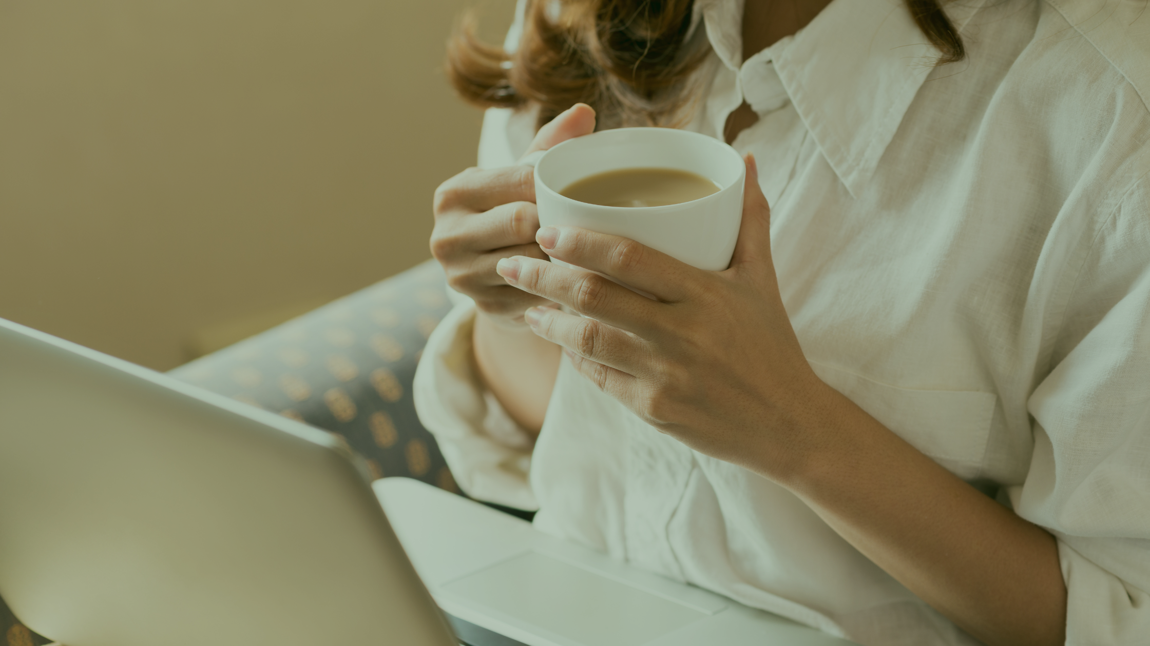 Person holding a white mug of hot coffee or tea, sitting at a table with a laptop.