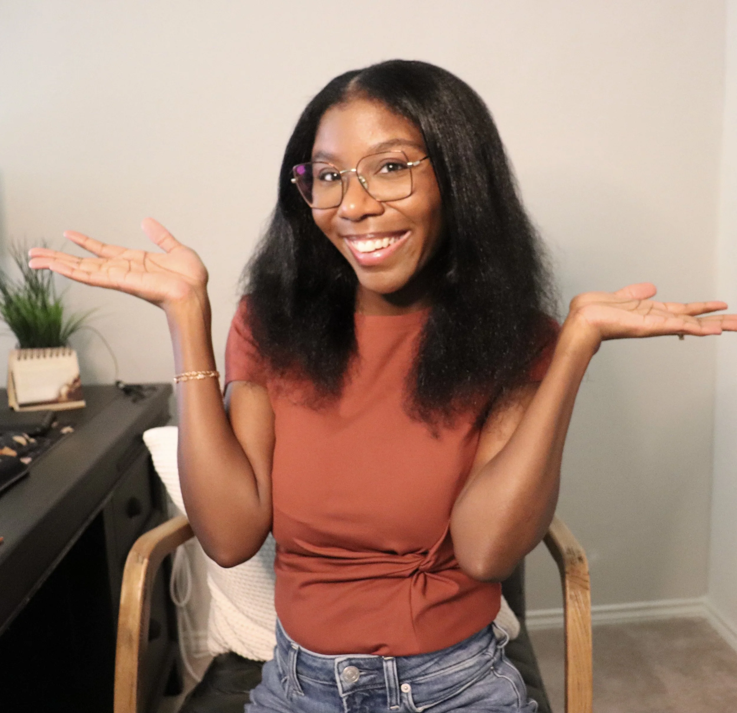 Close-up of a smiling woman with glasses, wearing a reddish-orange top and denim shorts, sitting on a wooden chair with arms raised, in front of a plain wall with a small plant to her left.