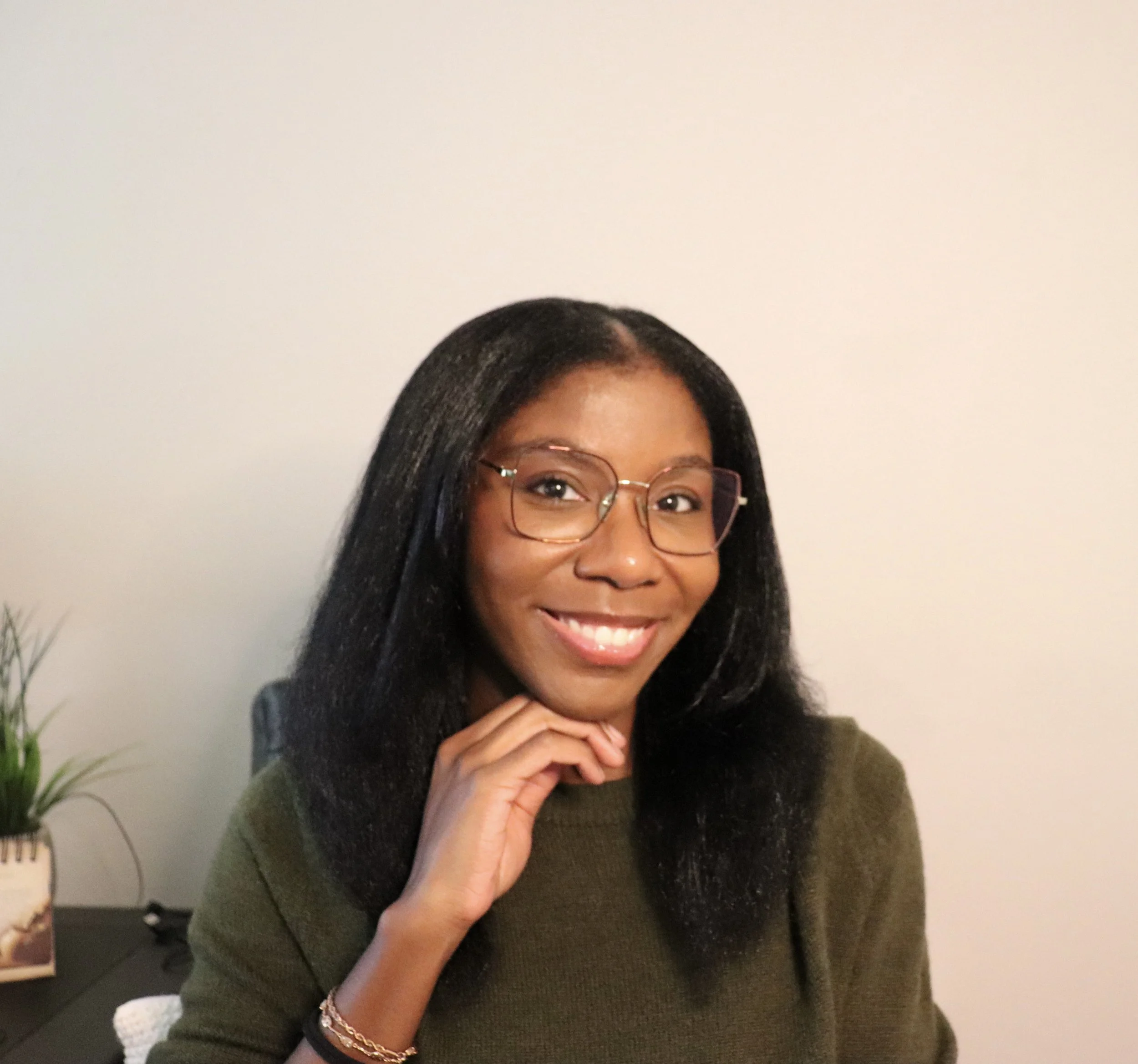 A woman with long black hair wearing glasses and a dark green sweater smiling and posed with one hand near her face in front of a light-colored wall.