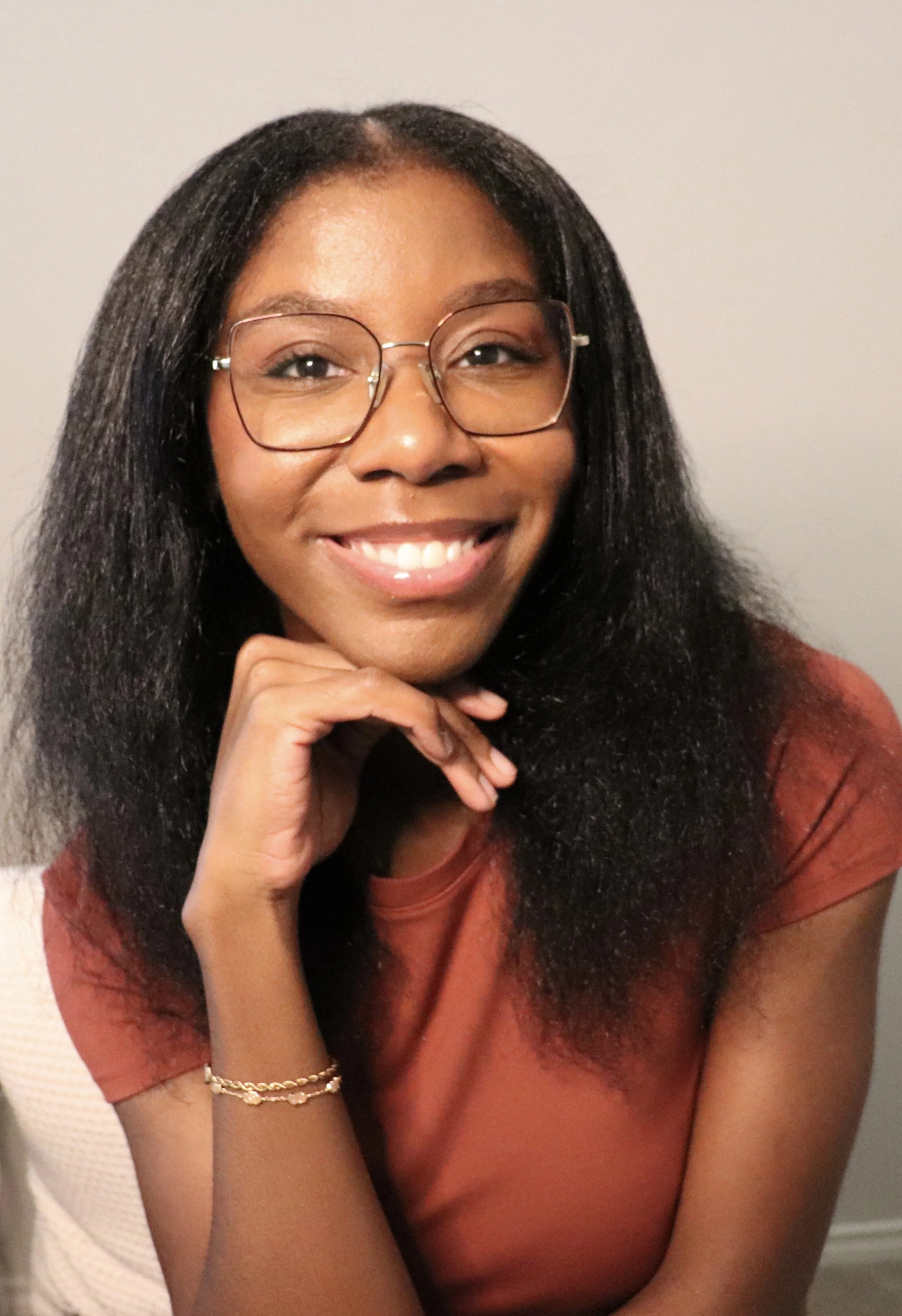 A young woman with glasses and long black hair smiling, resting her chin on her hand, wearing an orange shirt and a gold bracelet.
