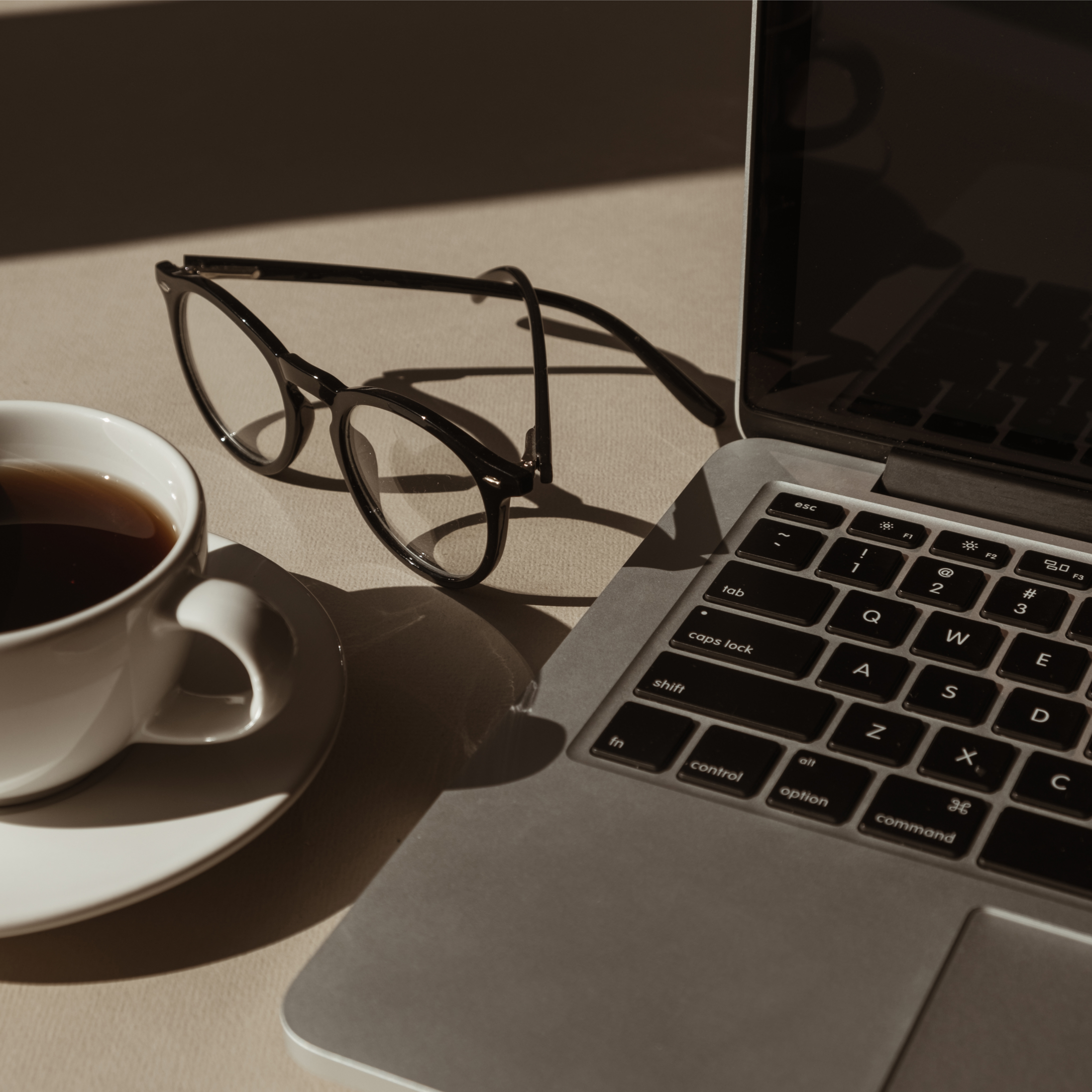 Close-up of a workspace with a coffee cup, black glasses, a laptop, and part of a keyboard on a desk.