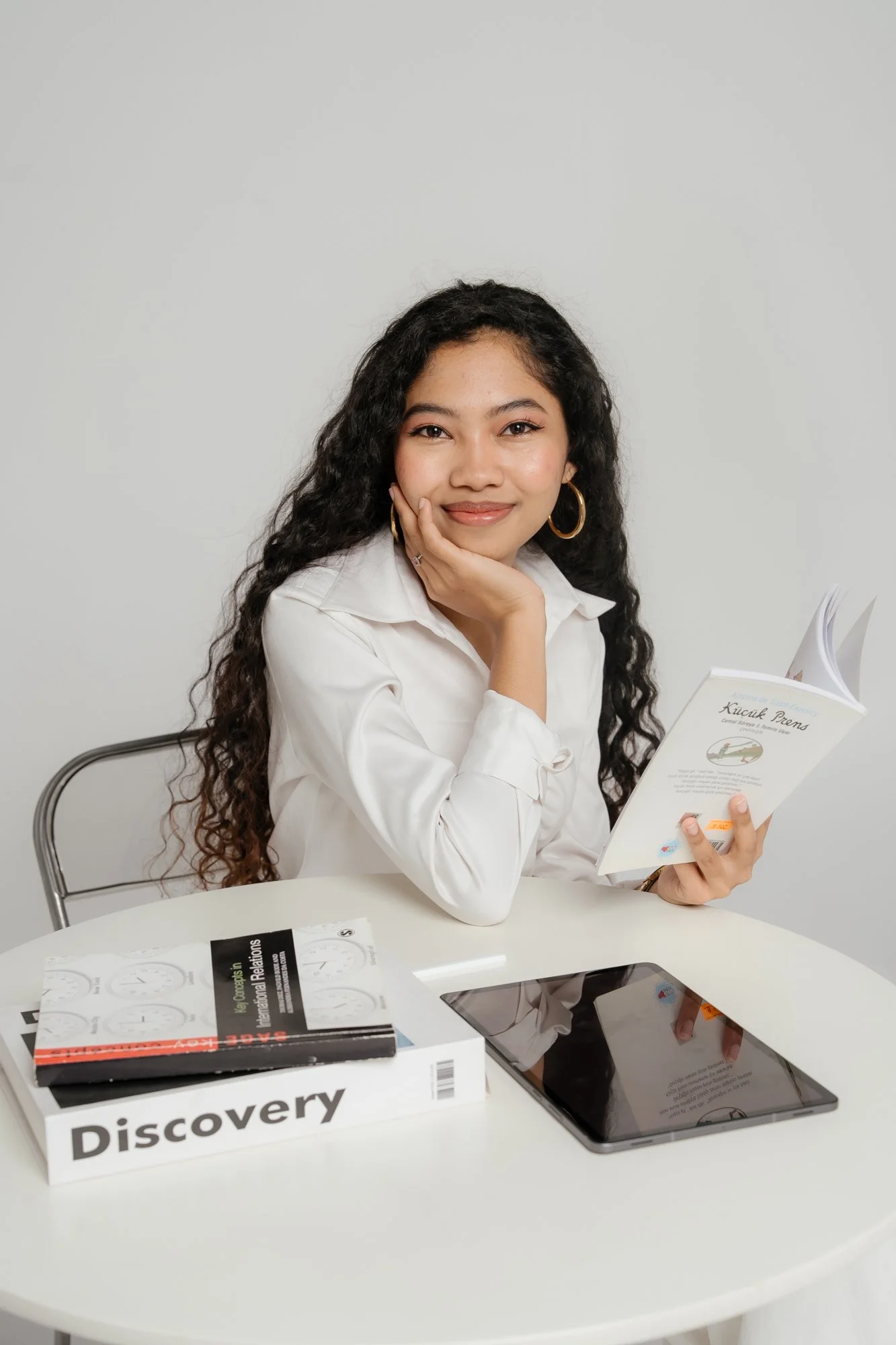 A woman with curly dark hair, wearing a white blouse and large earrings, sitting at a white table with books, a tablet, and a pamphlet, smiling and resting her face on her hand.