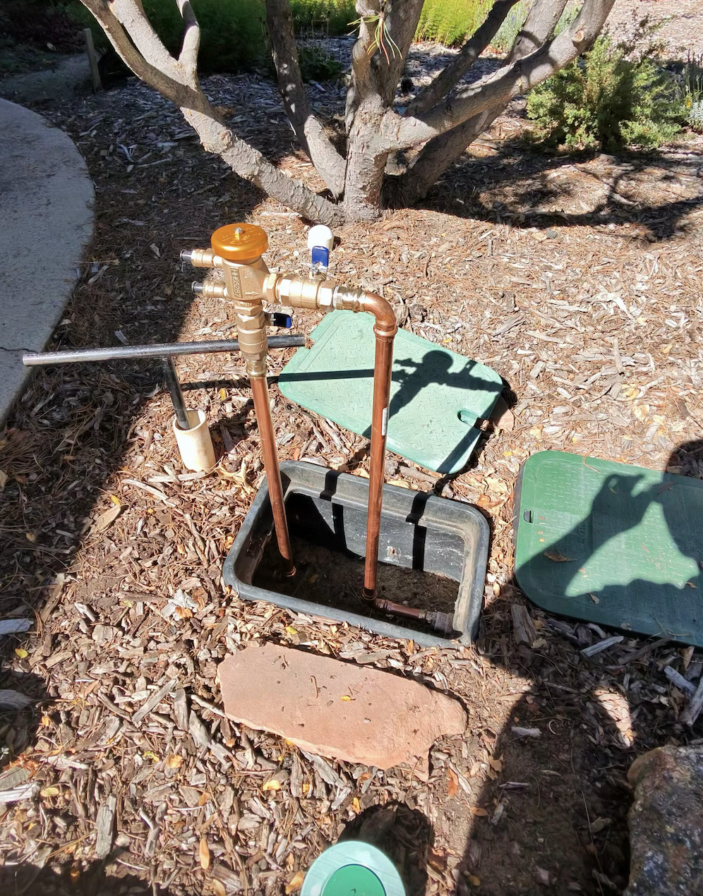 An underground water meter installation with copper pipes, a valve, and a green cover in a backyard with mulch, trees, and shrubs.