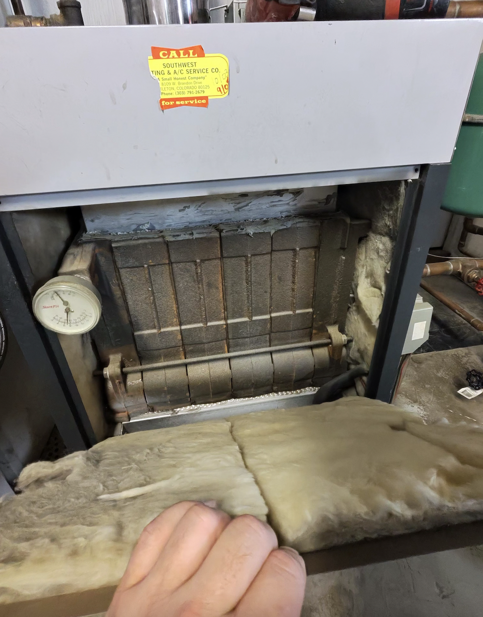 An HVAC system component with the front cover removed, revealing the internal coils and a pressure gauge, with insulation and tools nearby.