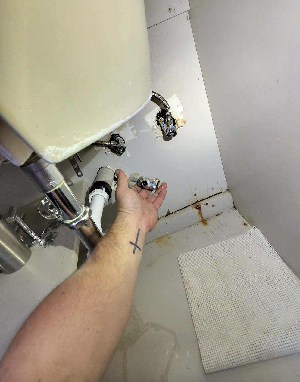 Close-up of a person's hand holding a damaged plumbing valve under a sink, with exposed pipes, rust stains on the wall and floor, and a white mat on the floor.