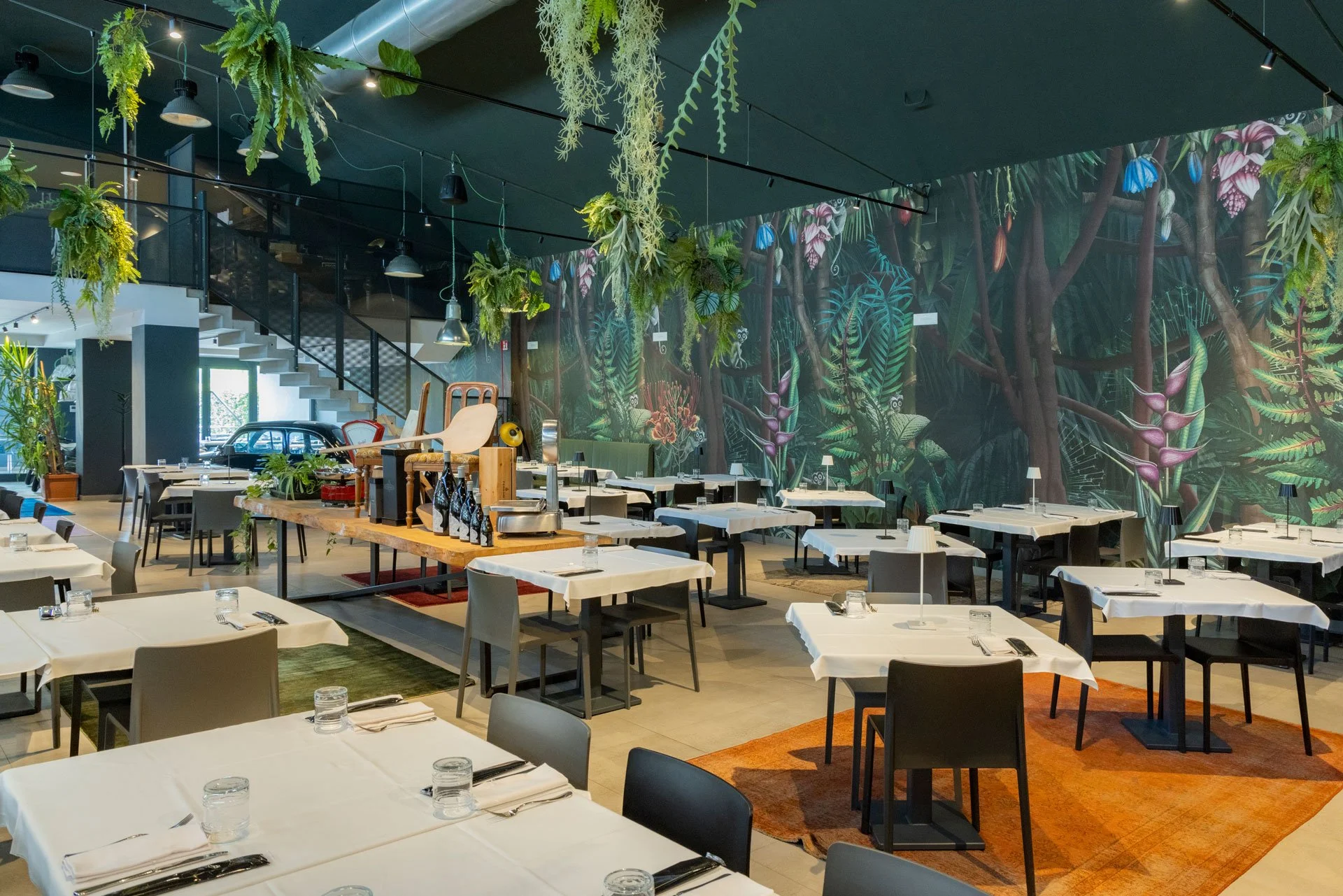 Sala da pranzo di un ristorante elegante con tavoli apparecchiati, piante sospese e murale di foresta tropicale