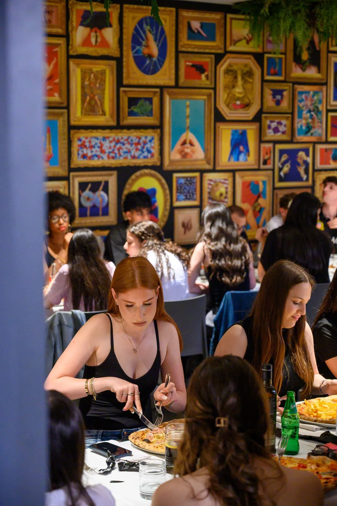 Ristorante affollato con persone che mangiano pizza e una parete decorata da numerosi quadri colorati