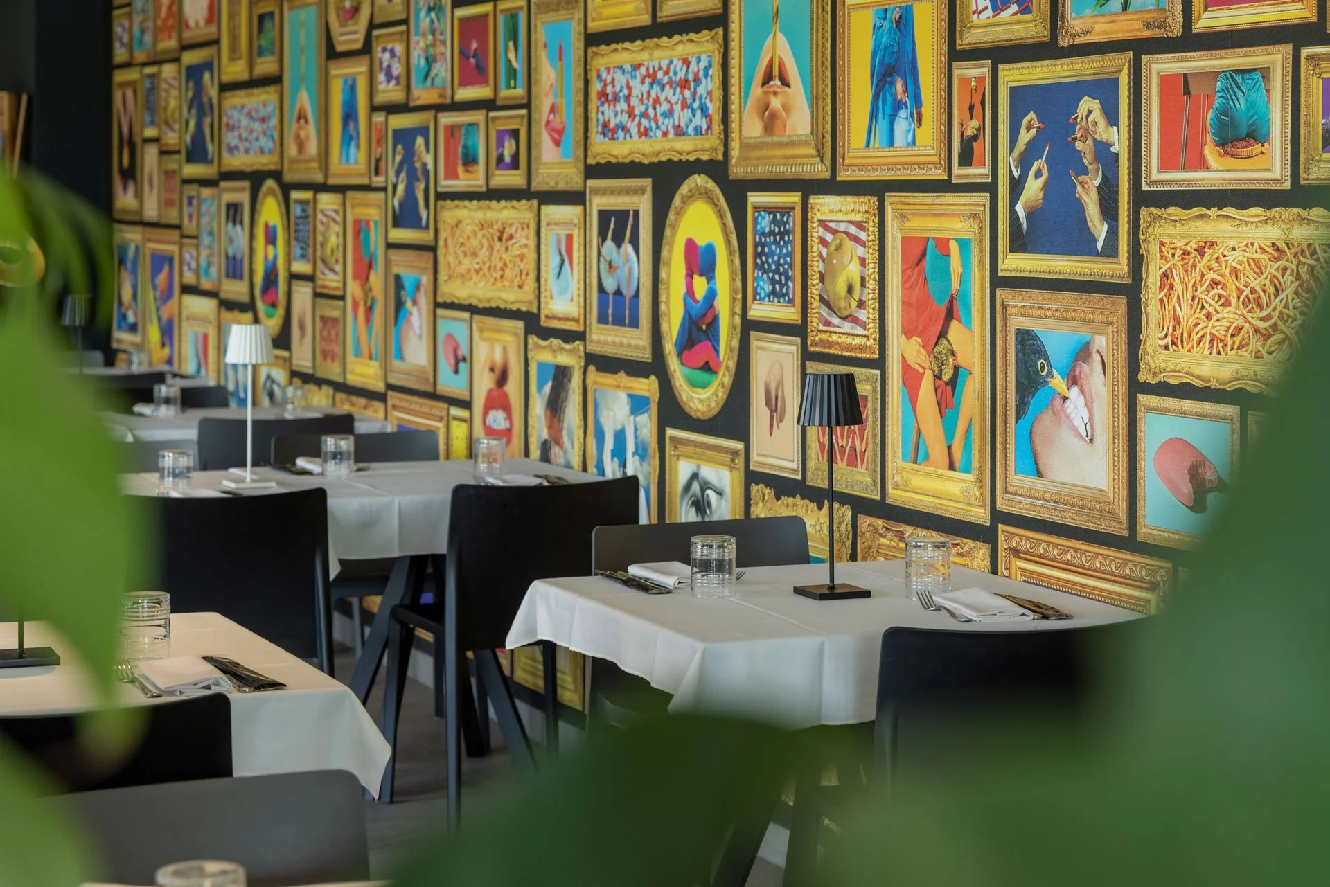 Sala di ristorante elegante con tavoli apparecchiati e parete decorata da numerosi quadri colorati con cornici dorate
