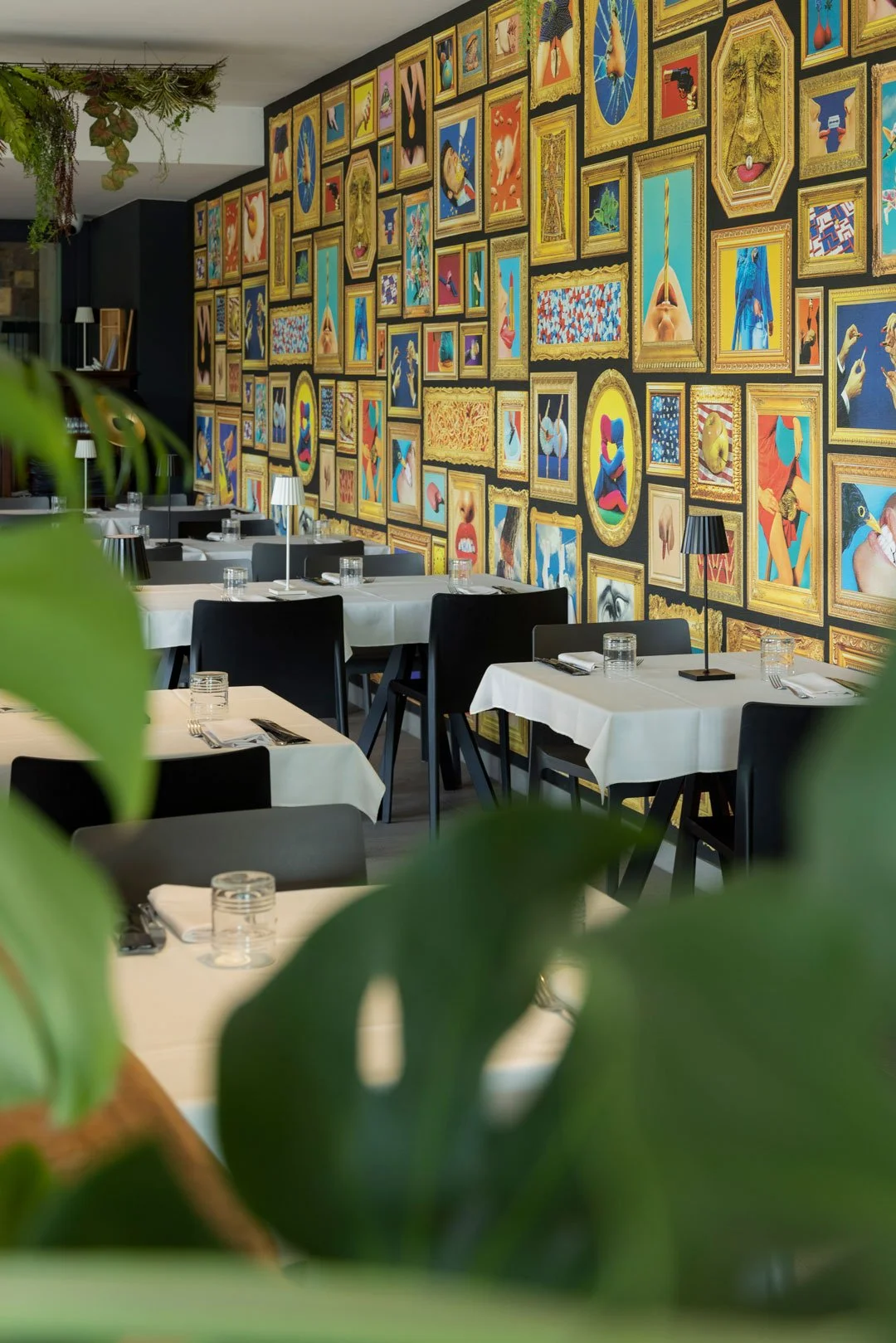Sala di ristorante elegante con tavoli apparecchiati, piante verdi e parete decorata da numerosi quadri colorati con cornici dorate.