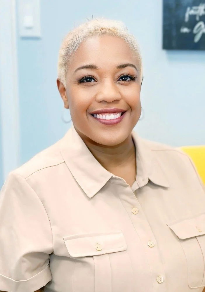 A woman with short blonde hair and a light beige collared shirt, smiling in an indoor setting with a light blue background.