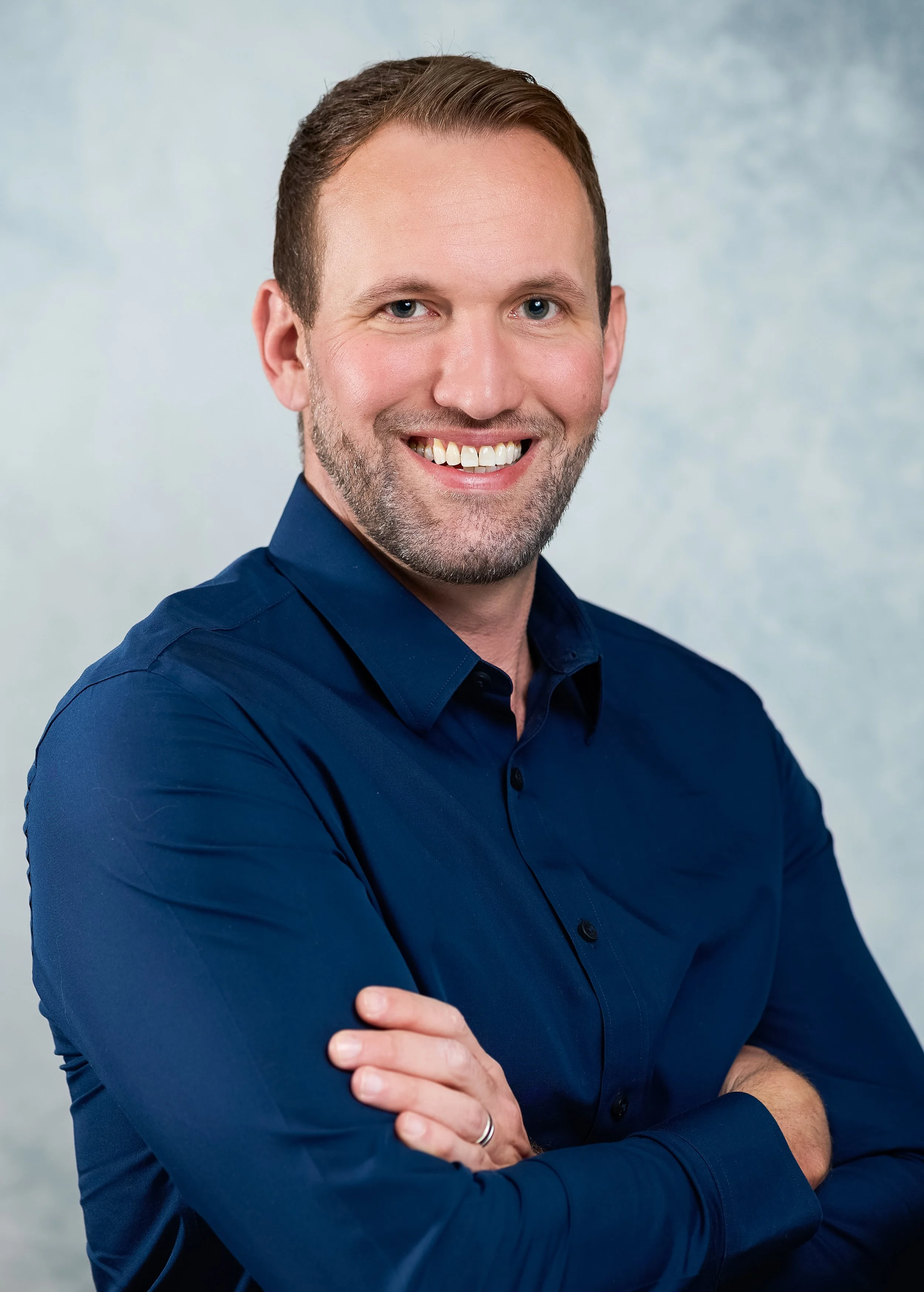 A man with short brown hair, blue eyes, and light beard wearing a dark blue button-up shirt, smiling, with arms crossed in front of a light blue background.