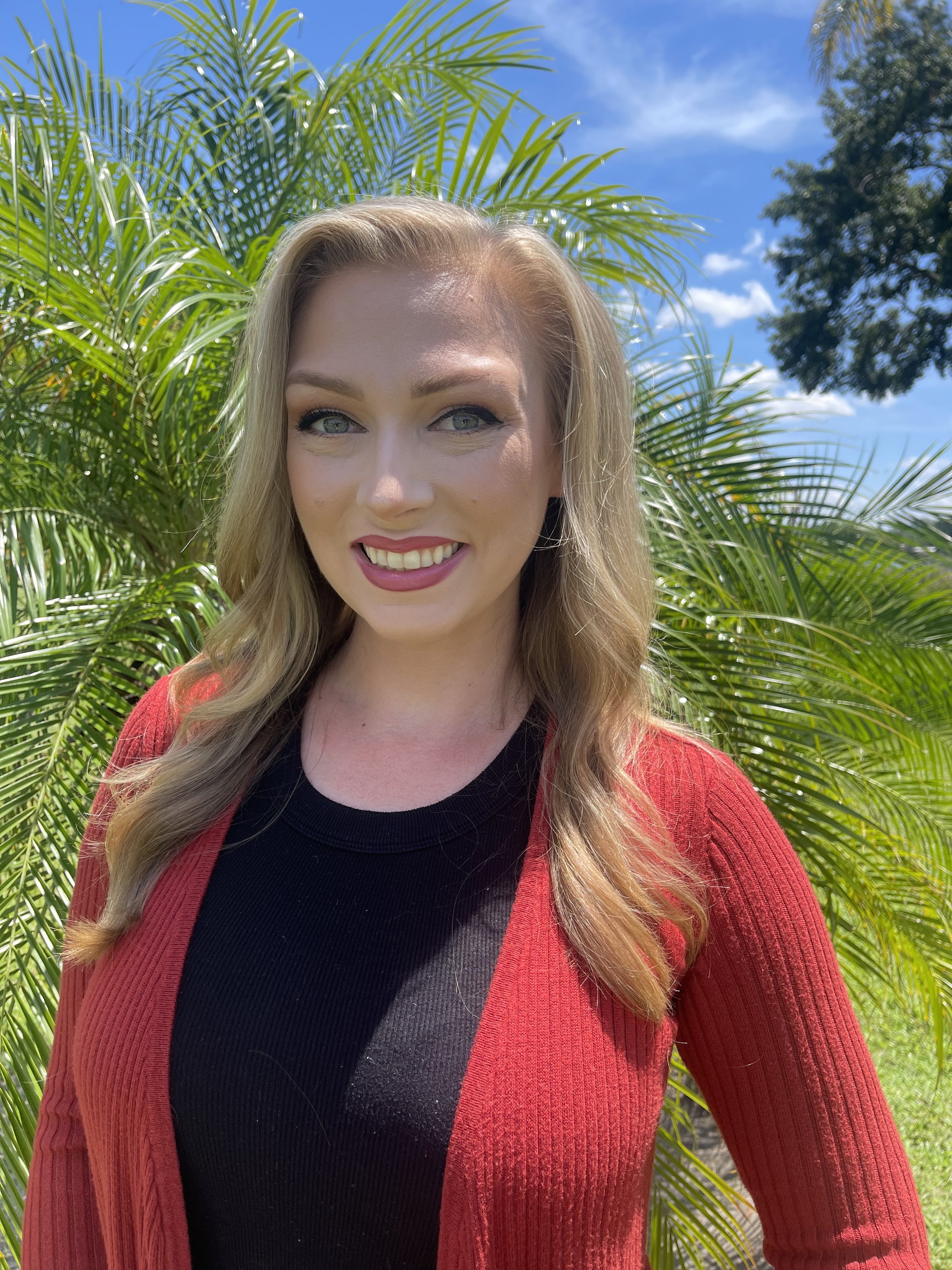 A woman with blonde hair and blue eyes smiling outdoors with a palm tree and clear blue sky in the background, wearing a red cardigan over a black top.