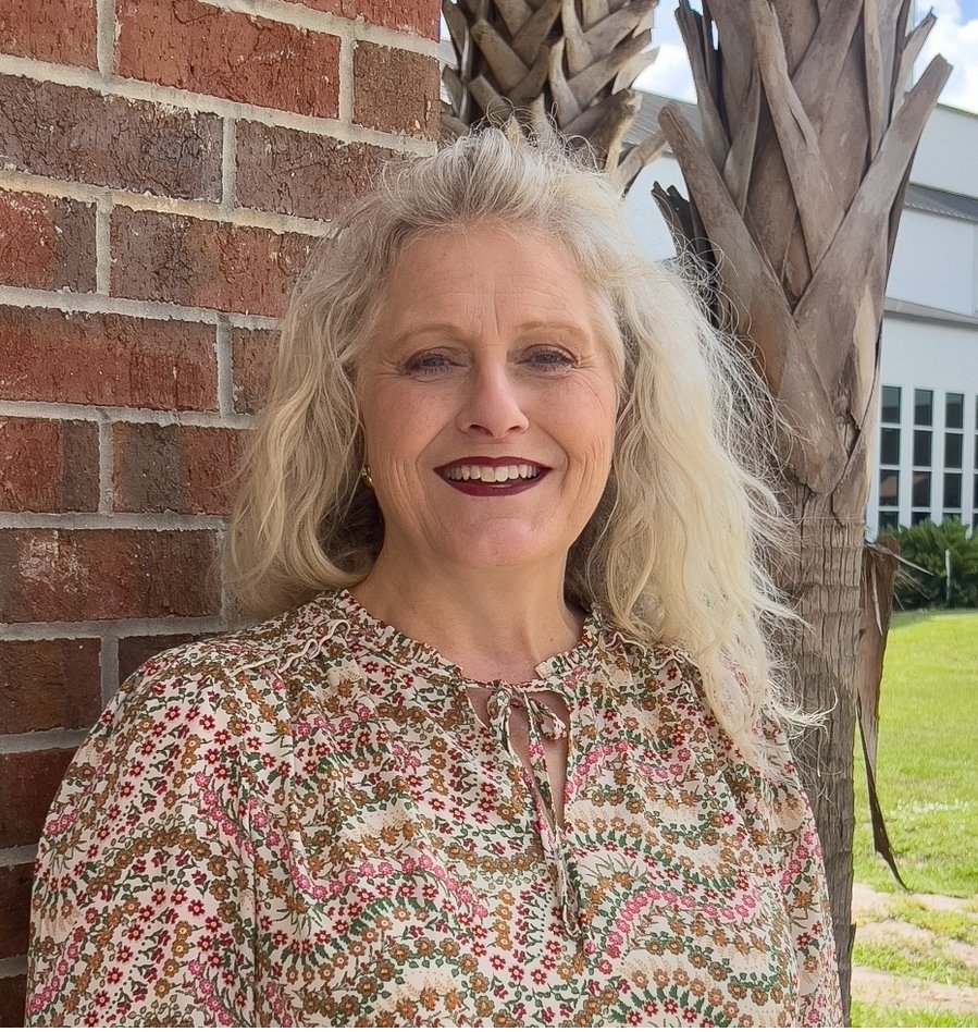 A woman with blonde curly hair smiling outdoors, wearing a patterned blouse, standing next to a palm tree and a brick wall.