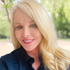 A woman with blonde hair and blue eyes smiling outdoors, wearing a dark blue top, with green trees in the background.