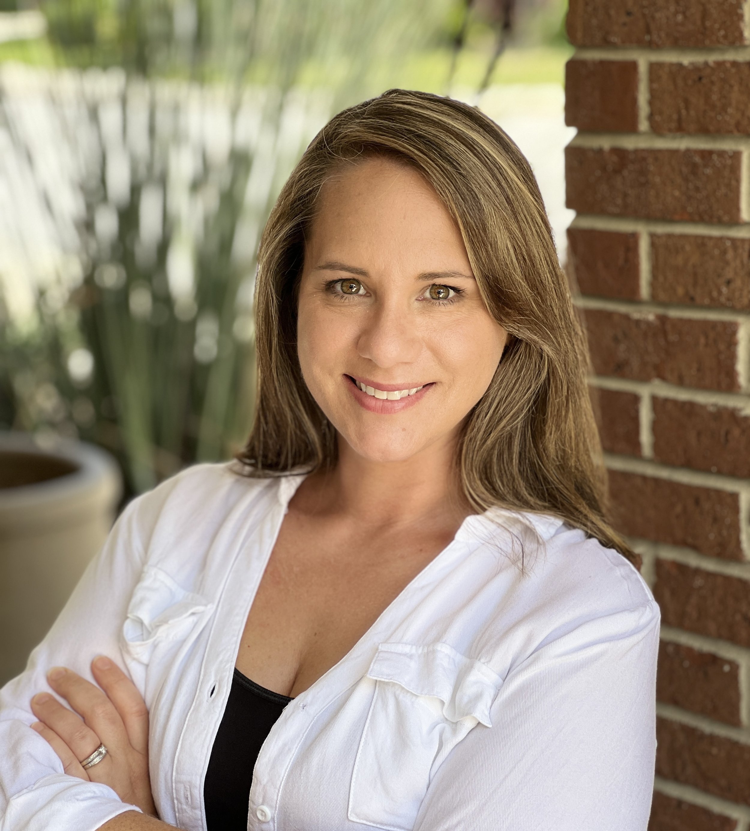 A smiling woman with brown hair and hazel eyes wearing a black top and white shirt, standing outdoors near a brick wall, with a potted plant and greenery in the blurred background.