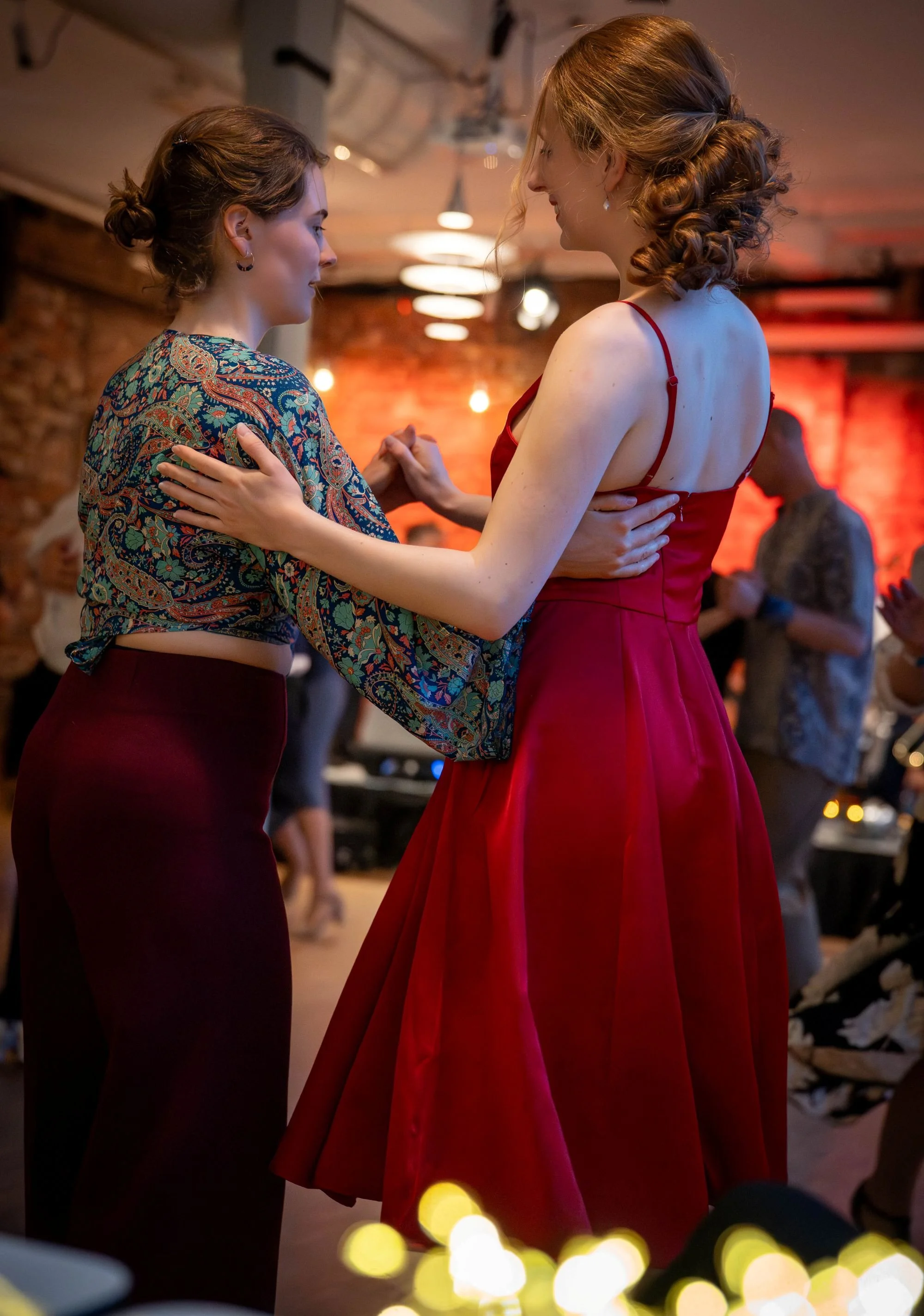 To kvinner danser sammen i et festlokale, kledd i formelle antrekk, med varmelamper og dekorerte vegger i bakgrunnen.