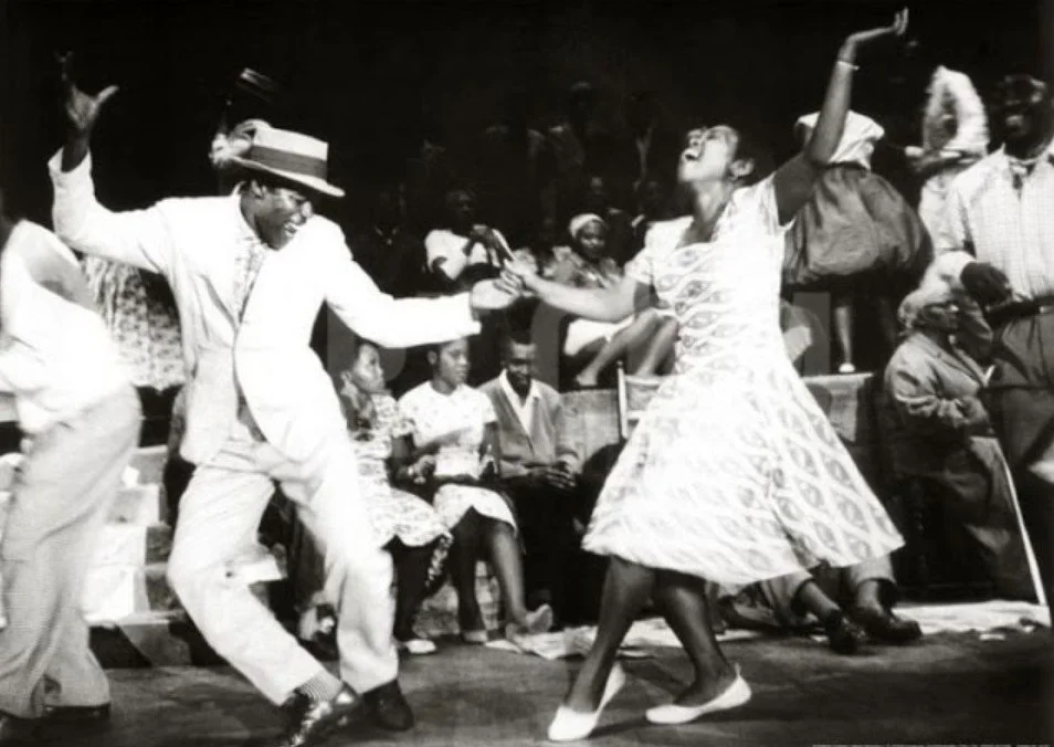 To personer som danser sammen, en mann i hvit dress og hatt og en kvinne i en mønstret kjole.
