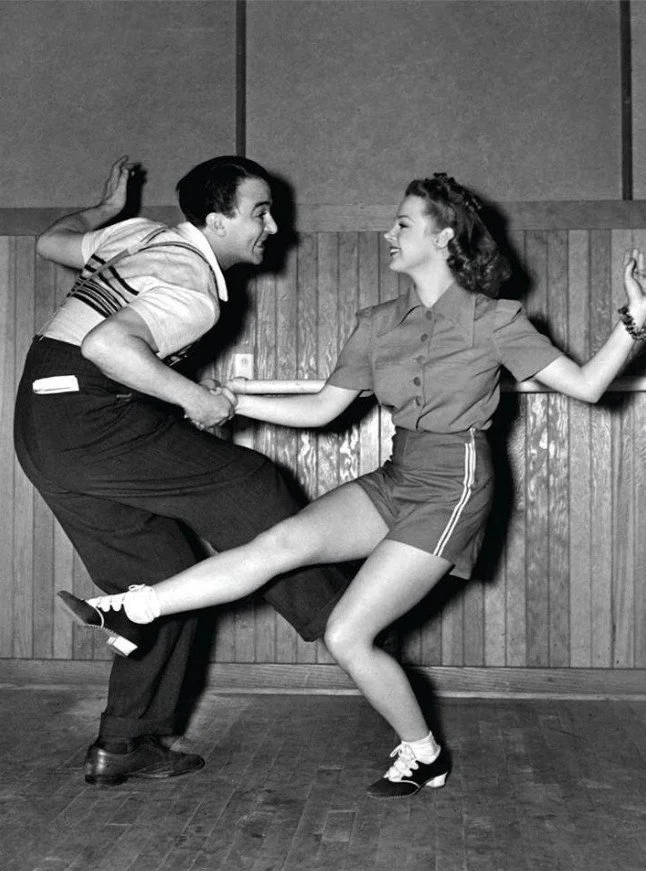 To personer danser i et rom med treffe vegger, den manlige danseren har på seg en stripet skjorte og mørke bukser, og den kvinnelige danseren har på seg en shorts og en knappestol bluse.
