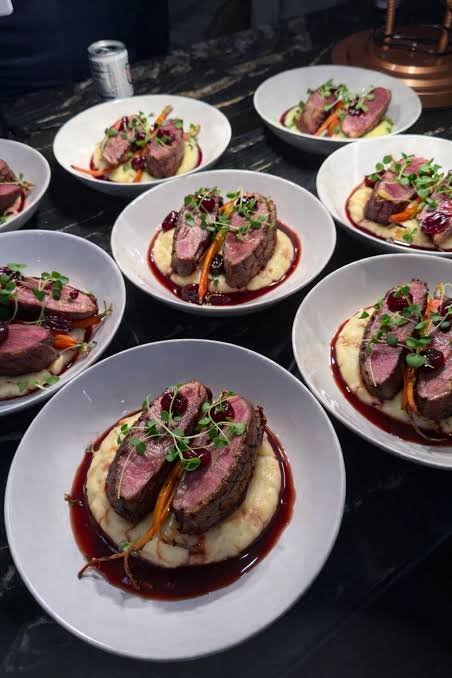 Six white plates of seared steak slices garnished with microgreens, served over mashed potatoes with a red wine reduction sauce, on a dark countertop.