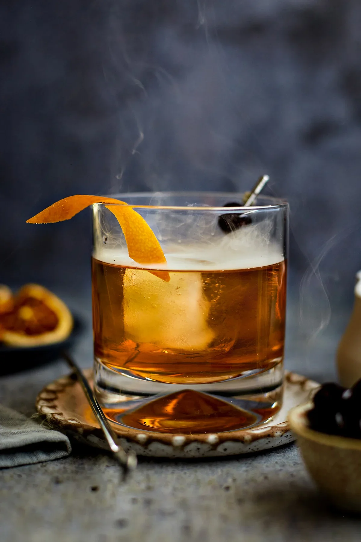 A glass of whiskey with an orange peel garnish and a straw on a textured surface, with dried citrus slices in the background.