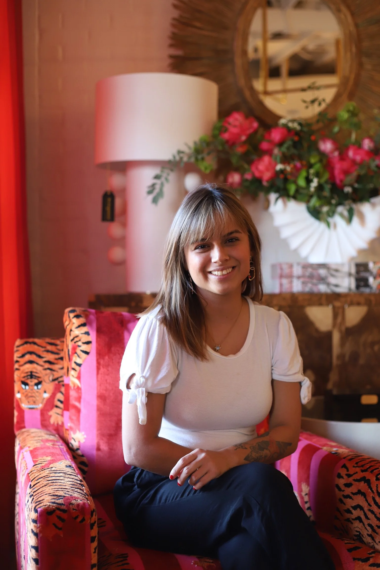 A young woman with shoulder-length light brown hair, wearing a white blouse with tied-up sleeves, smiling and sitting on a pink tiger-print chair in a cozy decorated room with a large bouquet of pink roses, a round mirror, a pink lamp, and colorful wall art.
