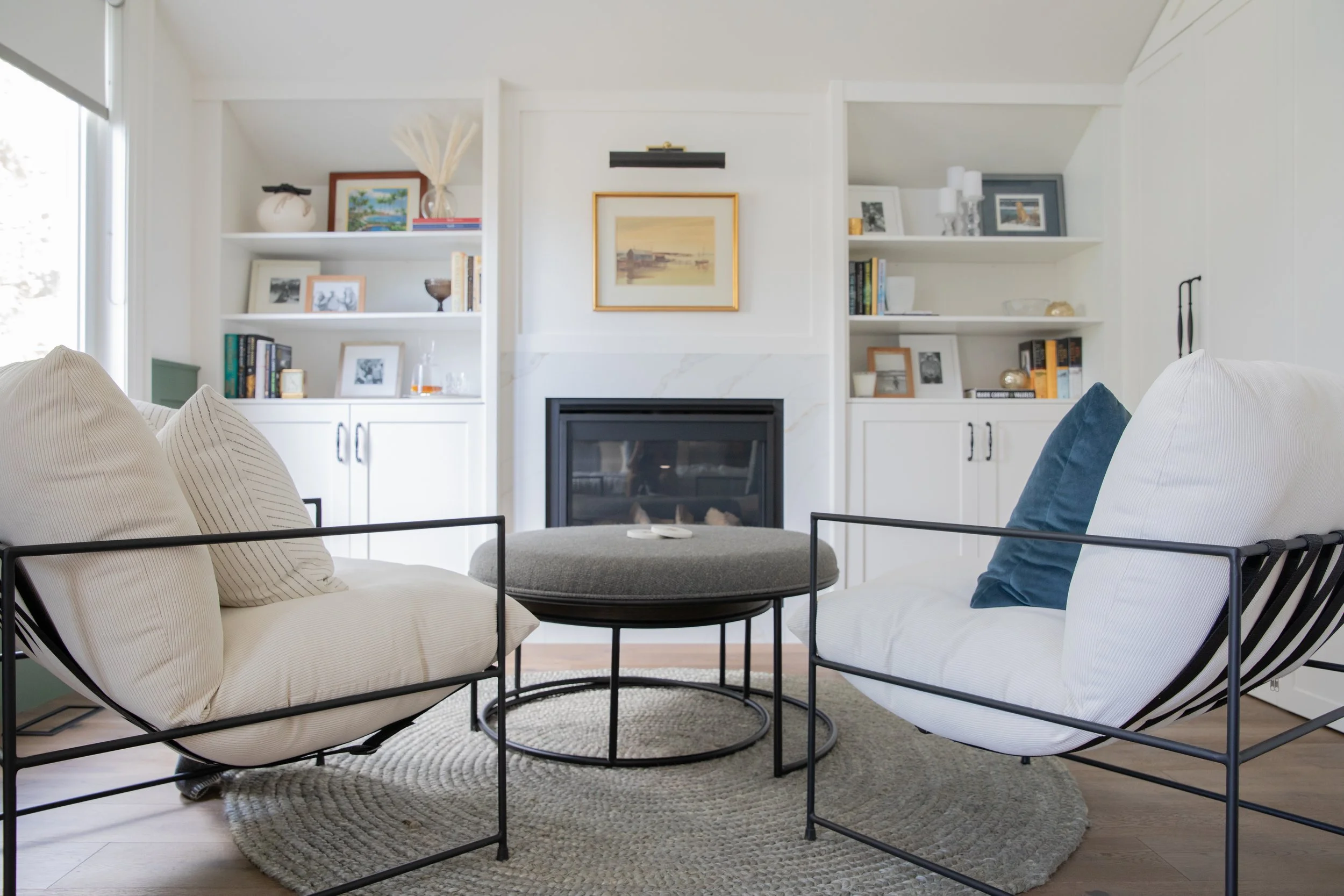A modern living room with white built-in bookshelves, a fireplace, and a dark metal coffee table with a round top. The couch is white with beige and dark blue pillows, and there's a round beige rug on the wooden floor. Framed art and decorative items are on the shelves.