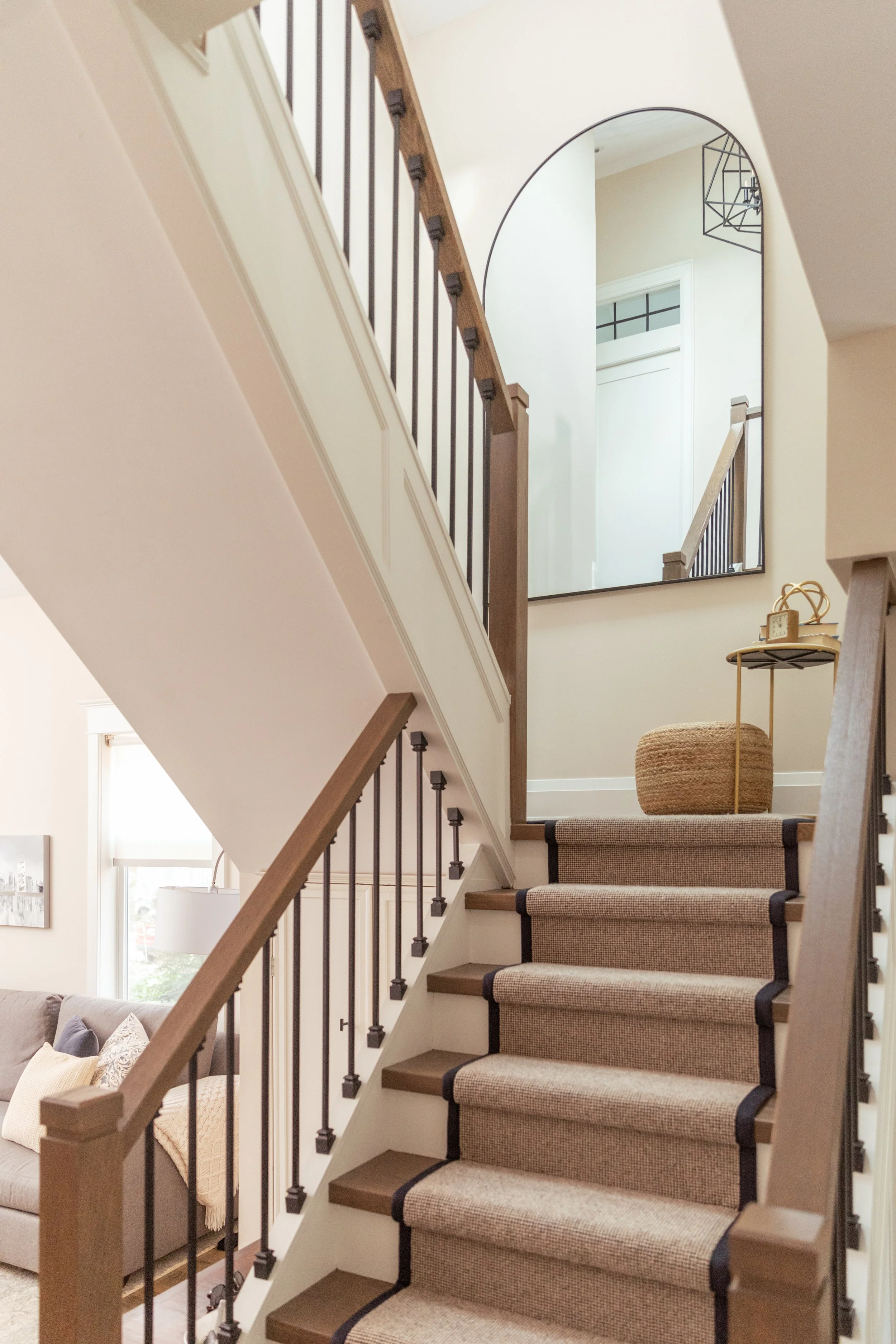 Interior staircase with beige carpet, wooden handrails, and black metal balusters, leading to an upper level with a large arched mirror reflecting part of the staircase and other interior elements.