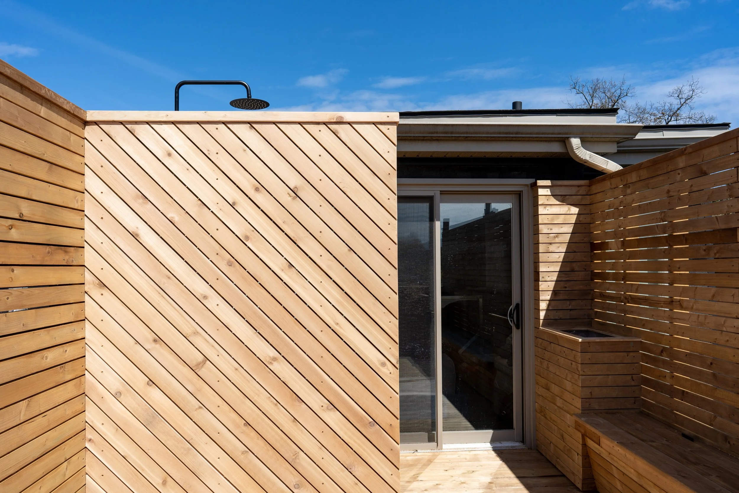 A modern outdoor balcony or terrace with wooden slat privacy walls, a glass sliding door, and a rain shower head visible against a clear blue sky.