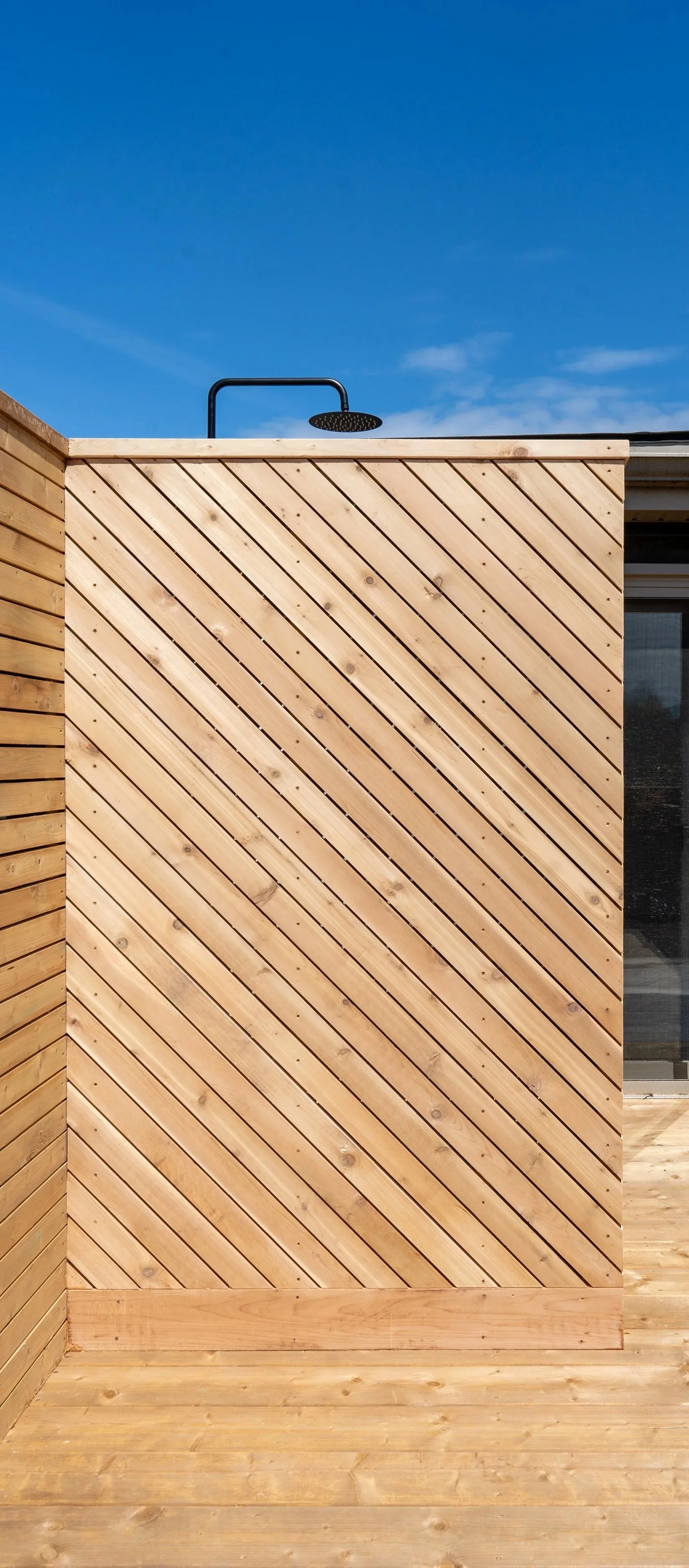 An outdoor shower area with made of wooden panels, a black showerhead, and clear blue sky in the background.