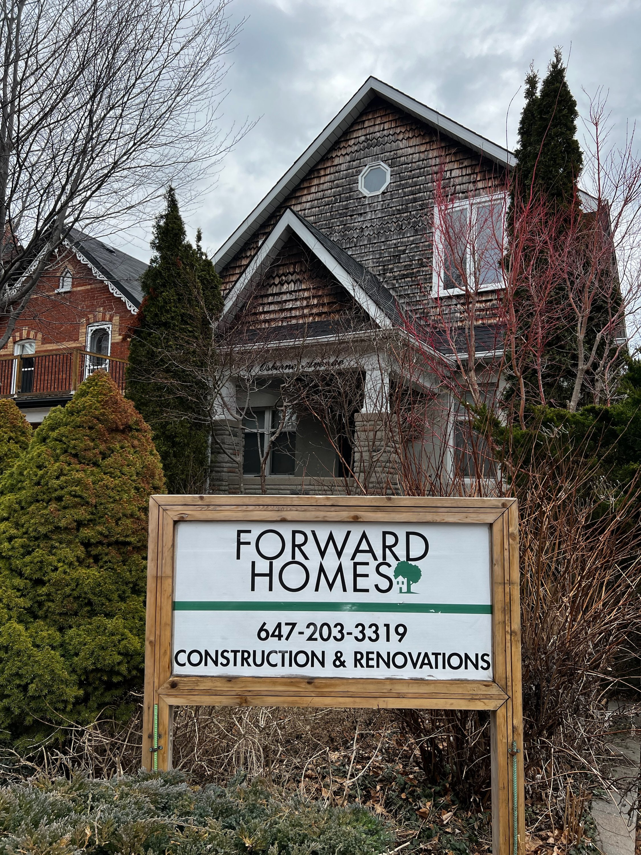 A house with wooden shingles, trees with no leaves, and a sign in front reading "Forward Homes" with contact information for construction and renovations.