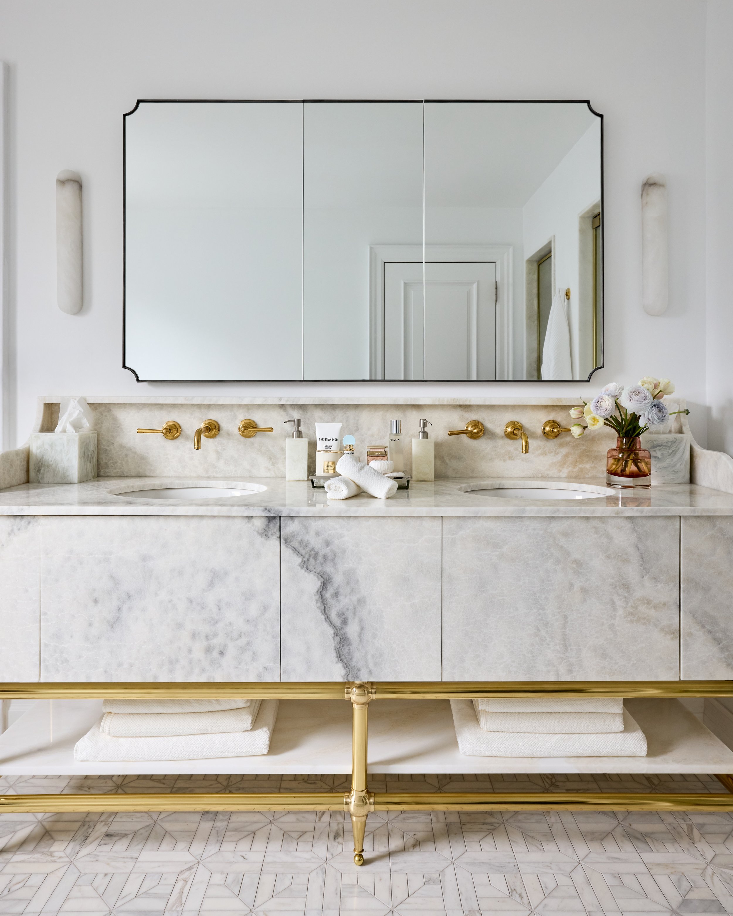 A luxurious bathroom vanity with a marble countertop, gold fixtures, a large mirror, and a vase of flowers.