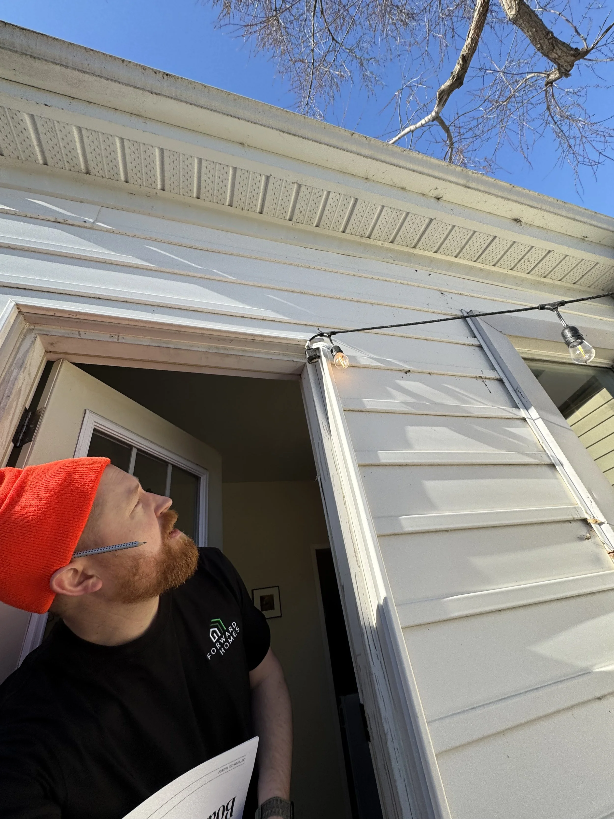 A man with red hair and a beard wearing a red beanie and black shirt, holding a document, standing outside a house with its door open. There are string lights with bulbs hanging from the house's roof, and a bare tree and blue sky in the background.