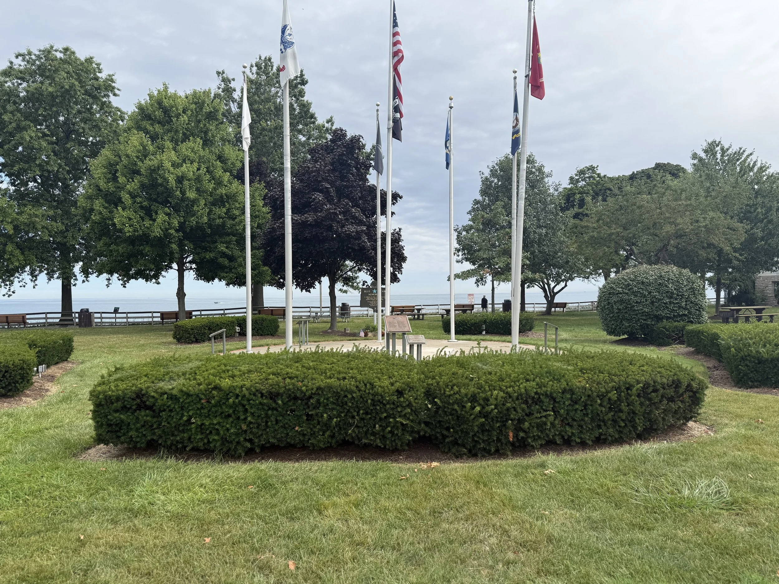 Park with a circular arrangement of nine flagpoles with various flags, surrounded by trees, benches, and shrubbery, with a view of the water in the background under a cloudy sky.