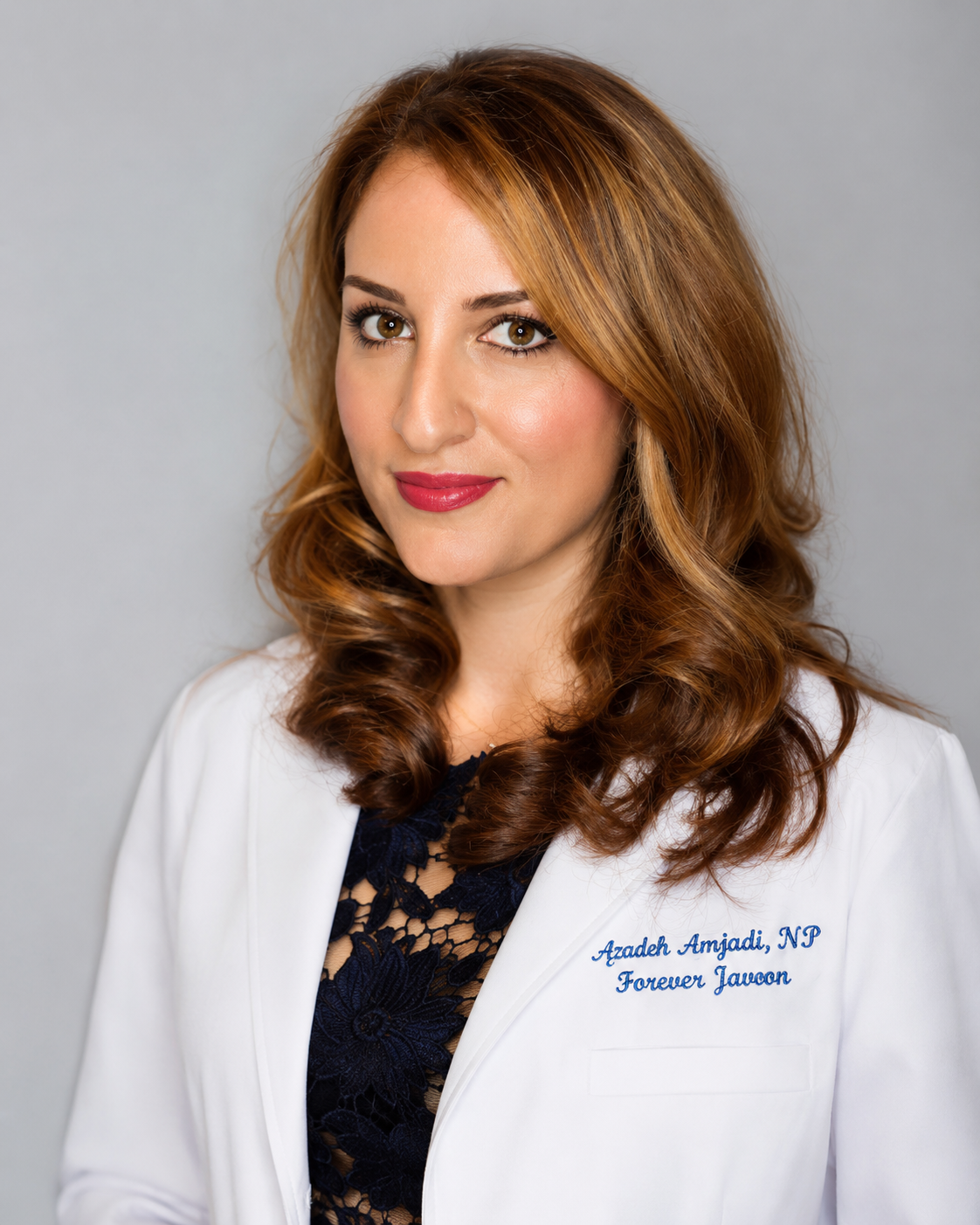 Portrait of a woman with red hair in a white coat with blue embroidery, smiling, against a gray background.
