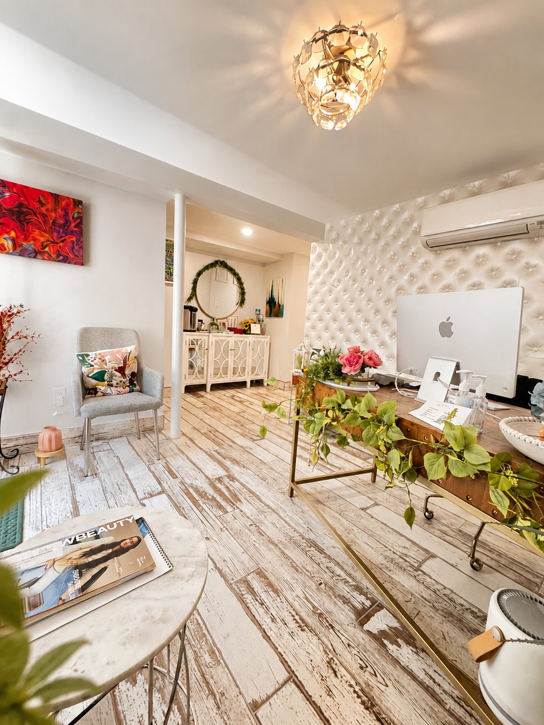 Interior of a cozy, modern living space with white walls, a tufted accent wall, a chandelier, a desk with an iMac decorated with plants and flowers, a cozy chair with a decorative pillow, a marble-top table with a magazine, and a small sideboard with artwork and a round mirror in the background.