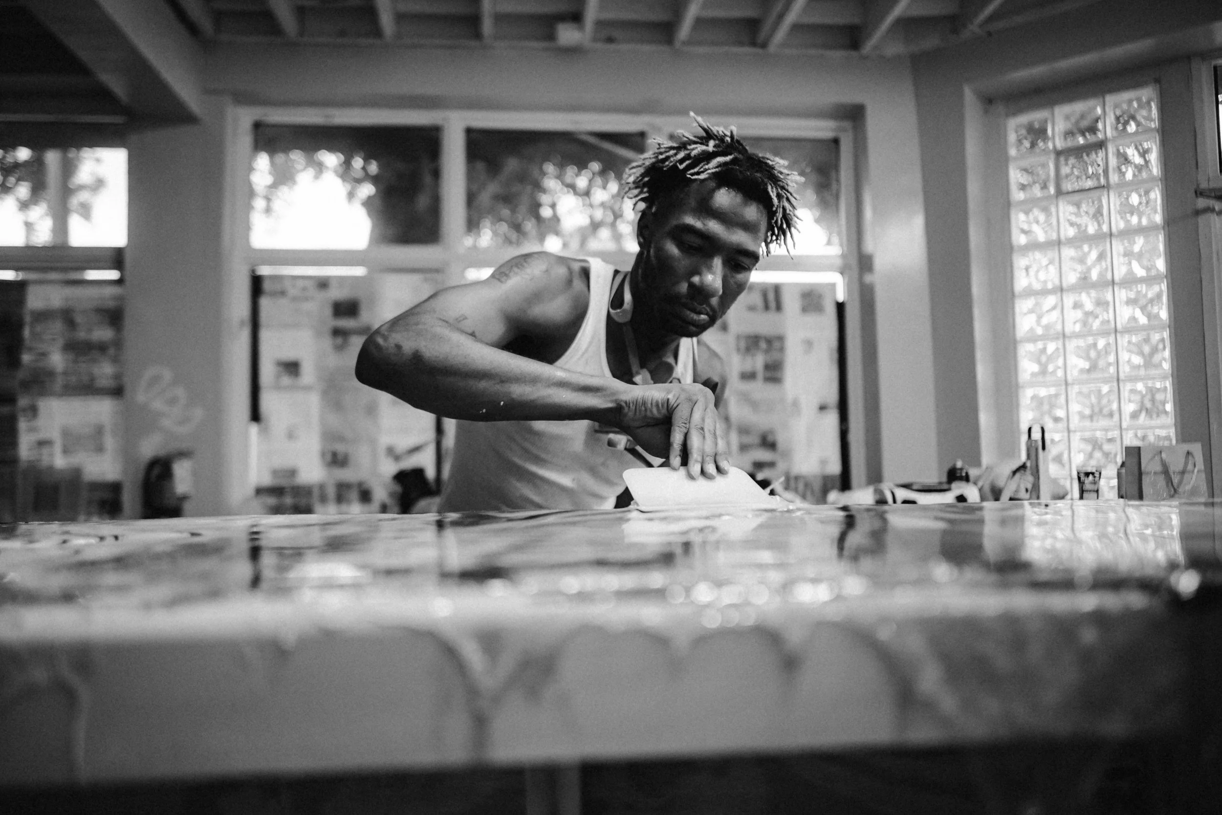 A man with dreadlocks in a sleeveless shirt leaning over a table with papers and objects, inside a well-lit room with large windows.
