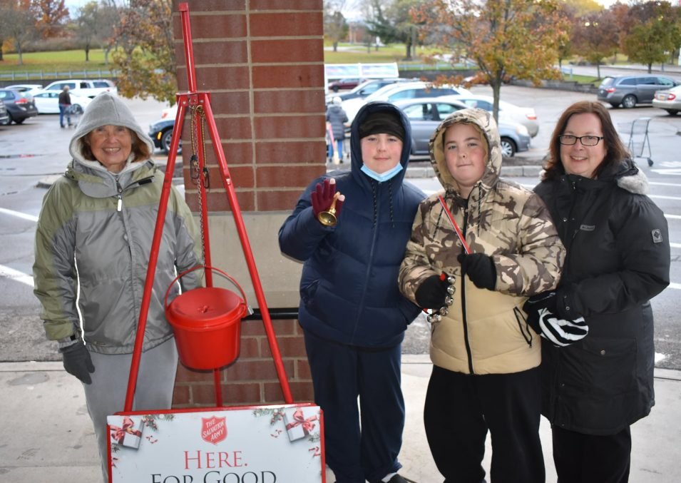 Two women and two boys standing outdoors in a parking lot with fall trees in the background. They are wearing jackets and gloves, and the boys are dressed in camouflage and dark winter gear. One boy is holding a bell, and there is a red kettle hanging on a tripod, with a sign that says 'HERE. FOR GOOD.' for a charity donation drive.