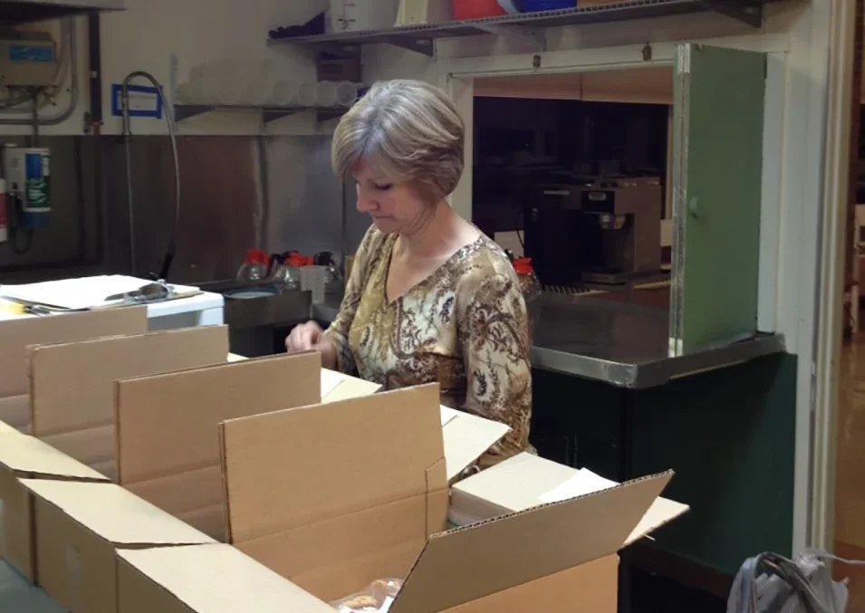 A woman with short gray hair wearing a patterned blouse working behind a box with divider sections in a kitchen-like setting with industrial equipment.
