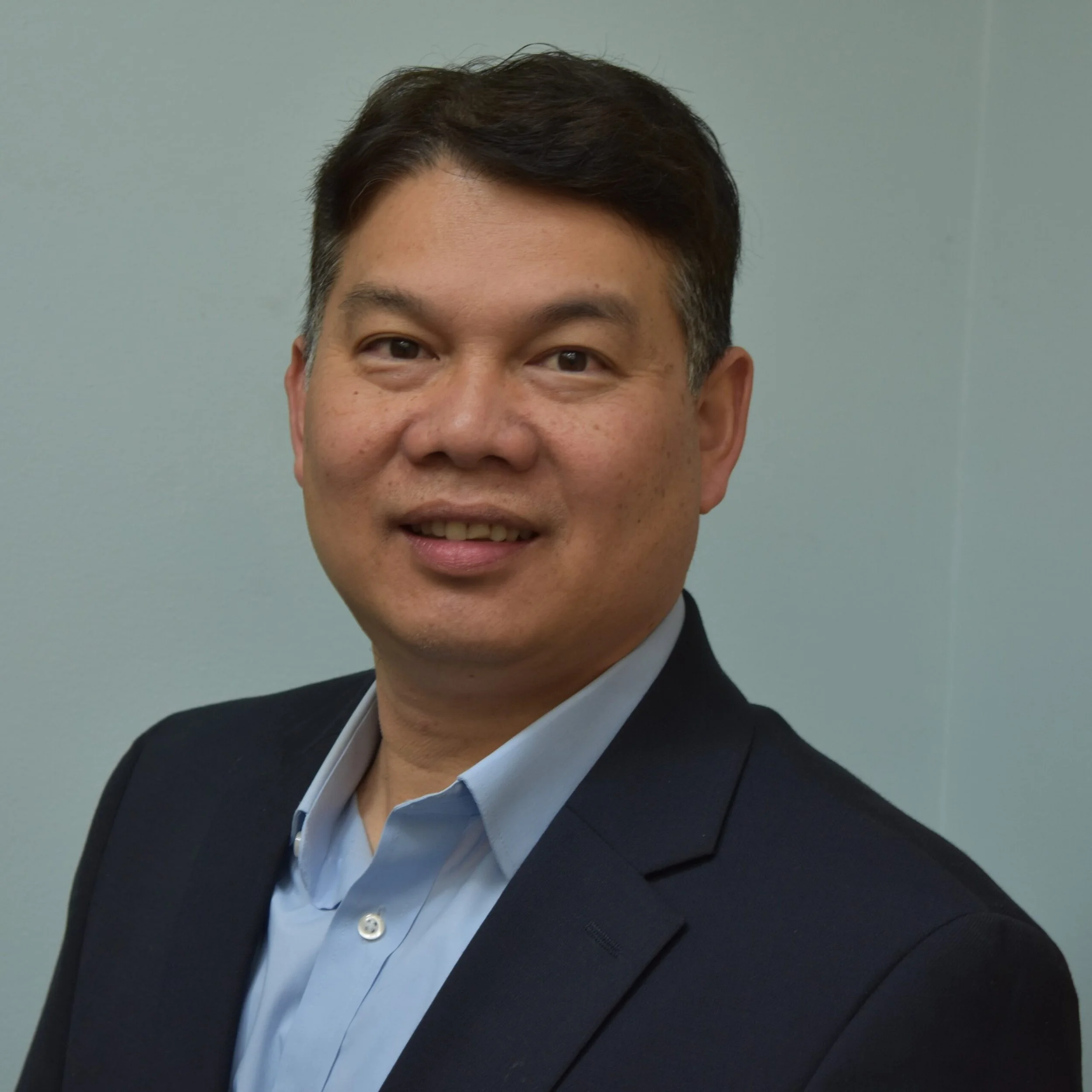 Close-up of an Asian man in formal attire, including a black blazer, sky blue shirt, and dark tie, smiling against a plain light gray background.