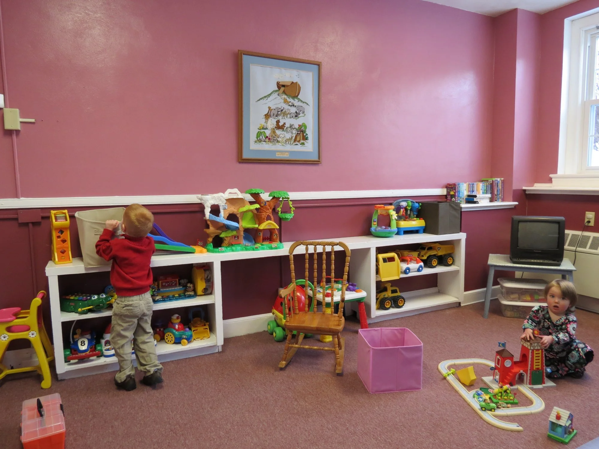 Children playing in a colorful nursery with pink walls, toys, and books. Service is streamed here live