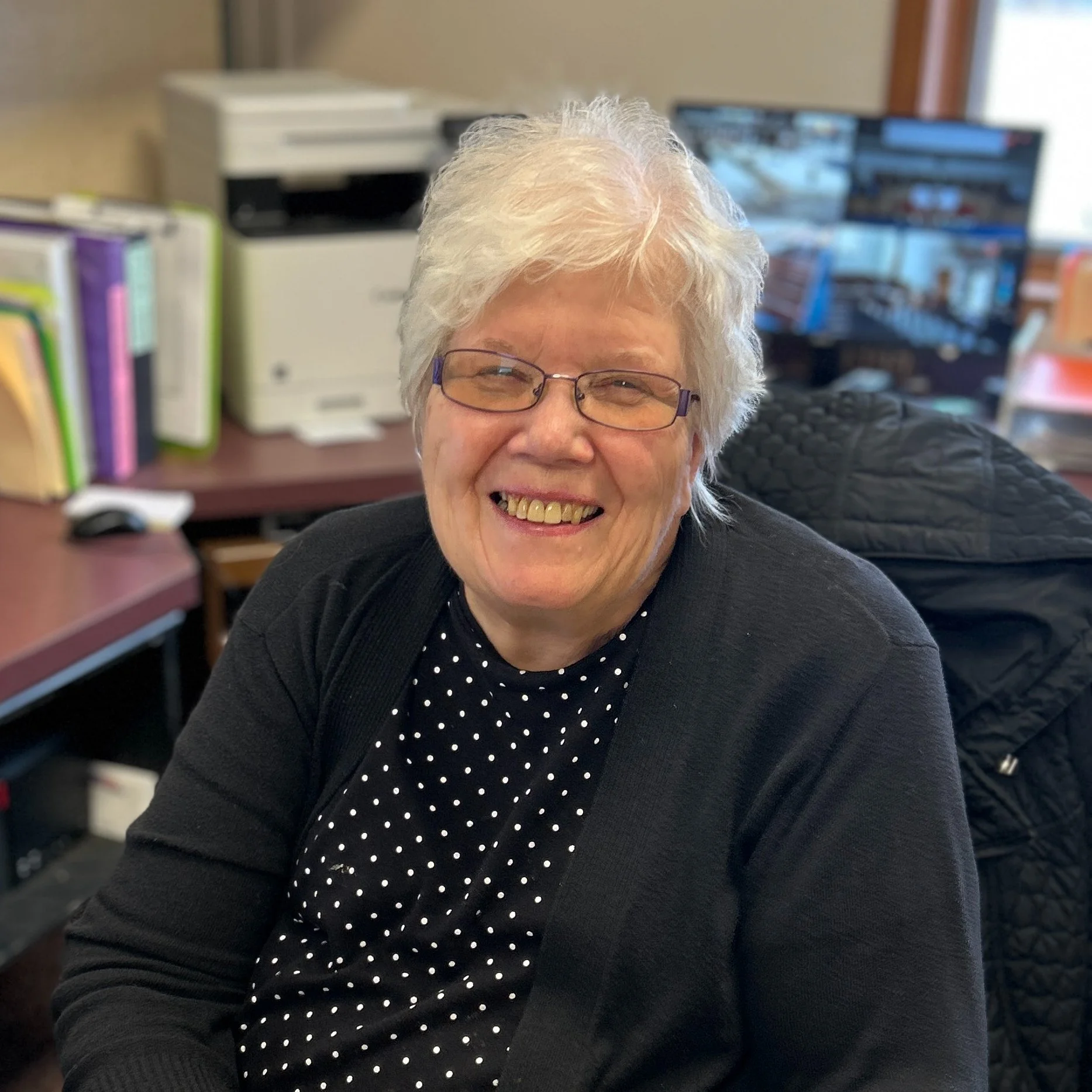 Close-up of a smiling elderly woman with short white hair and glasses, sitting at a desk with office supplies and a computer monitor in the background.