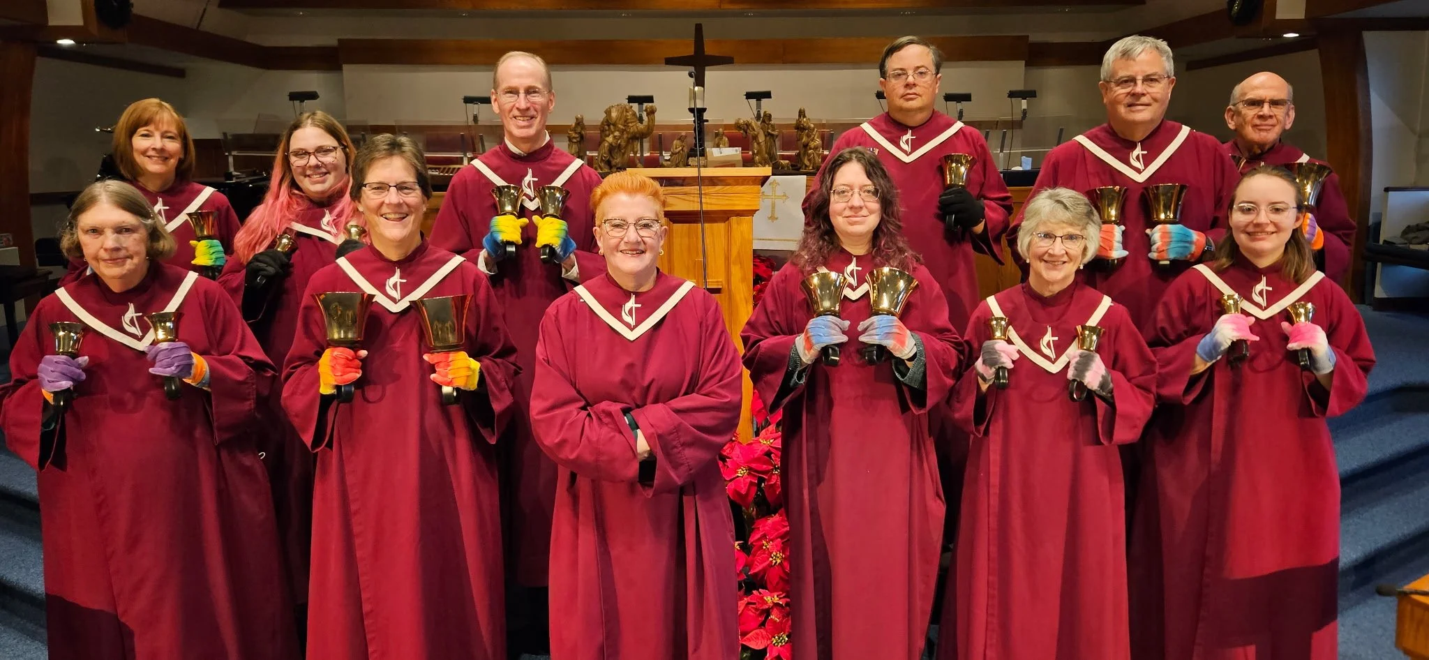 A group of fifteen people dressed in choir robes, holding bells, standing inside a church near a pulpit with religious statues and a cross in the background.