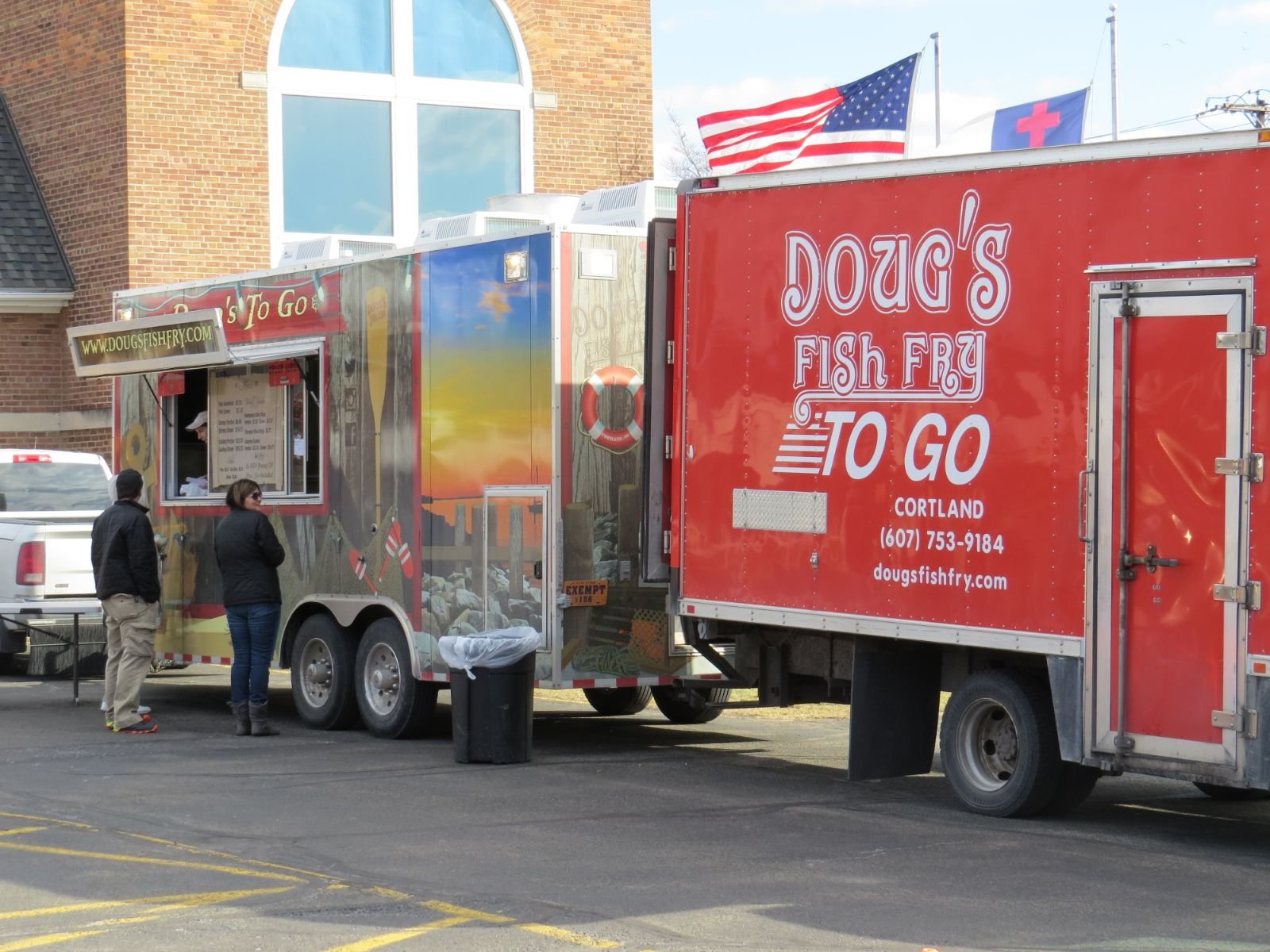 Red food truck with white text advertising fish and chips to go, parked in a parking lot near a brick building with large windows. Two people are standing near the truck, and flags of the United States and Red Cross are flying above it.