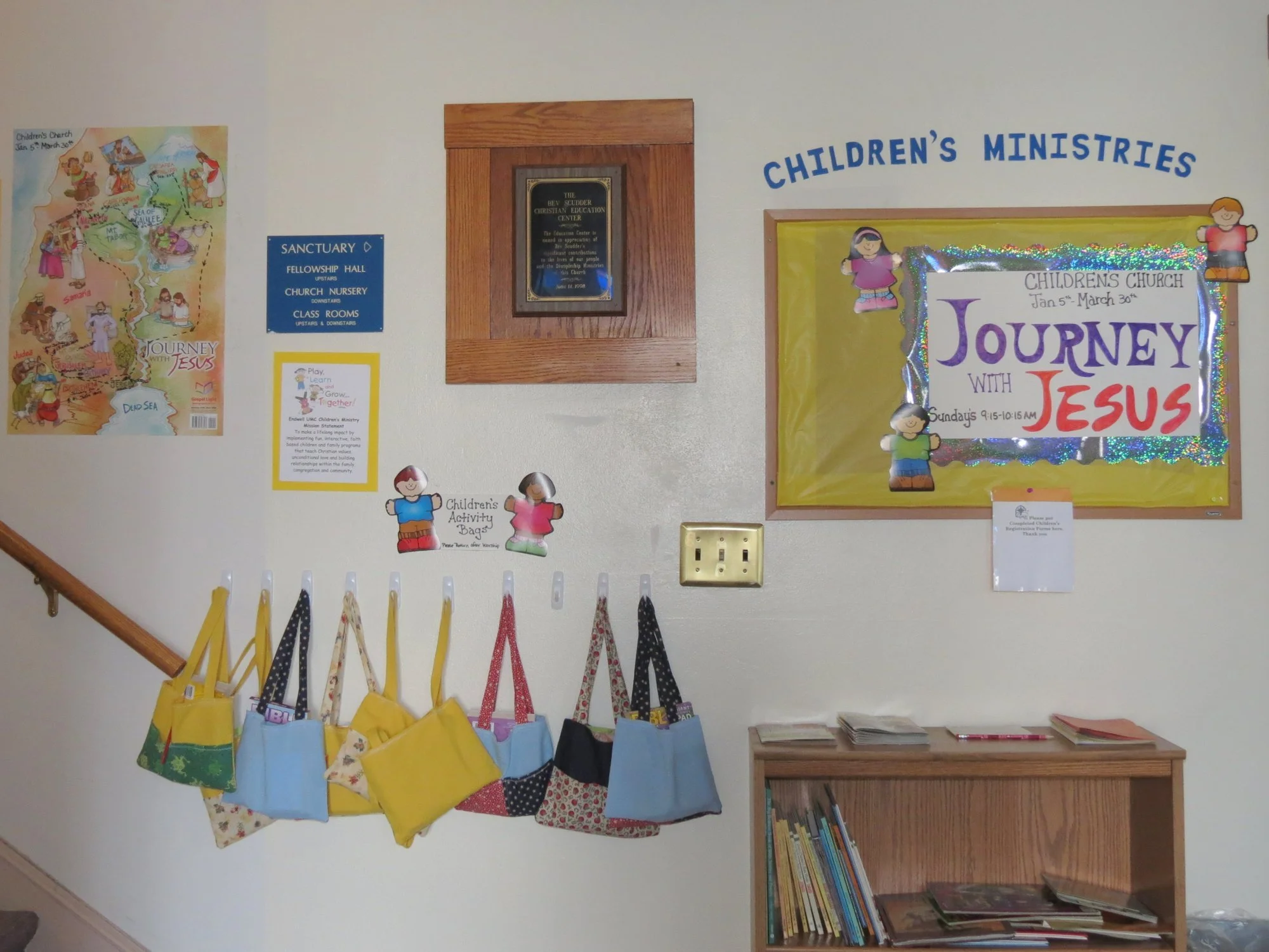 Wall with colorful children's church posters, hanging fabric bags, and a wooden bookshelf with books.