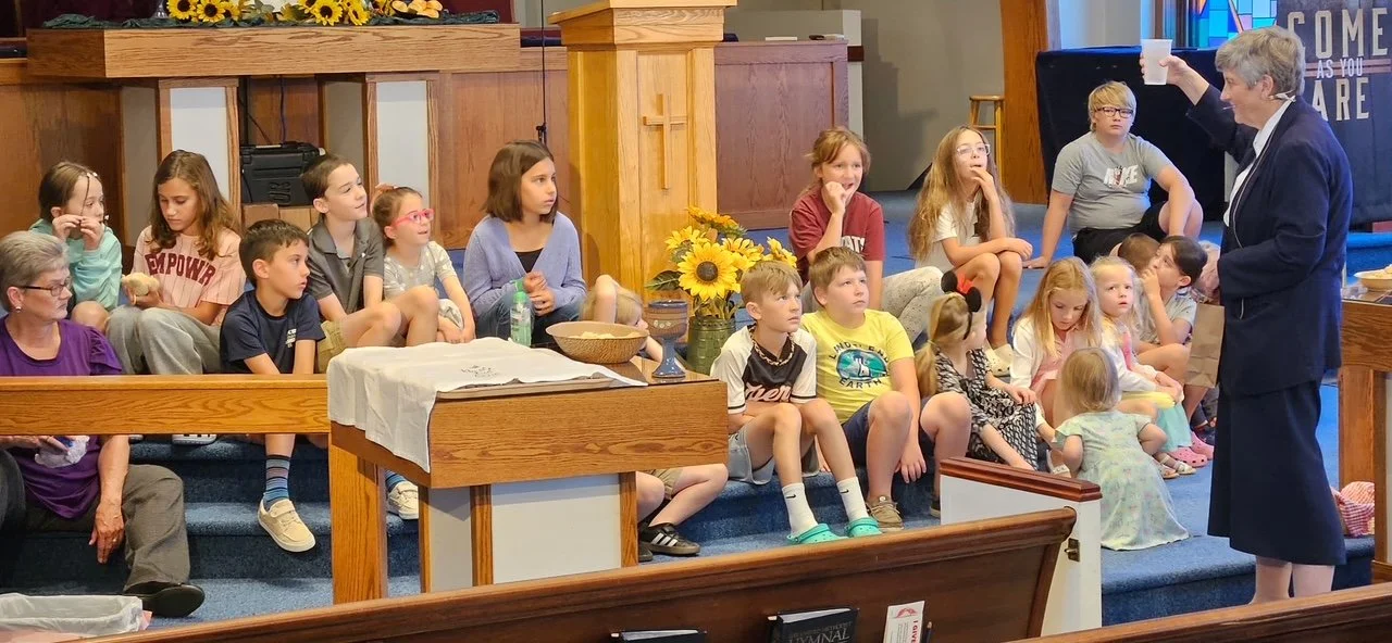 A woman in a suit is holding up a cup, speaking to a group of children and adults seated on church pews and stairs inside a church.