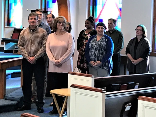A group of people standing inside a church near the altar, with stained glass windows in the background.