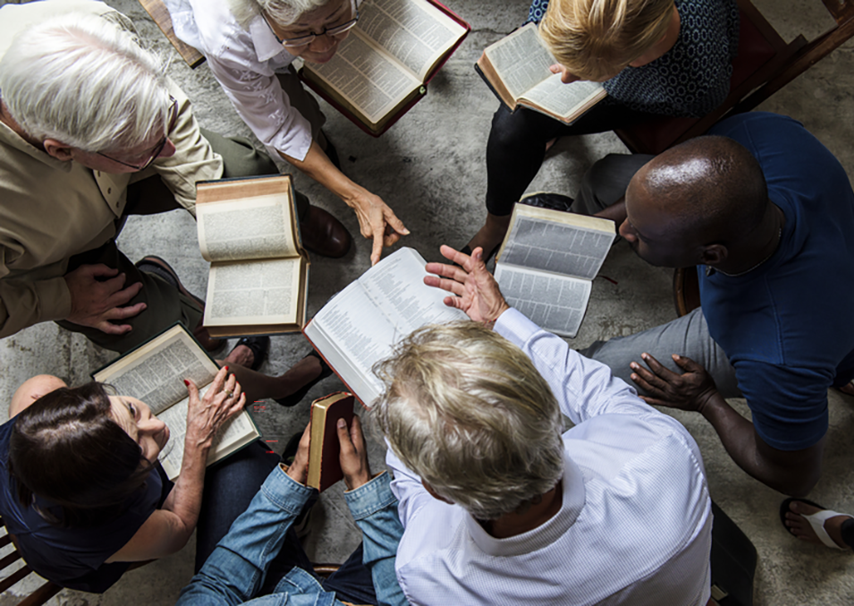 A diverse group of six people gathered in a circle, reading and discussing from open books in a room with a concrete or stone floor.