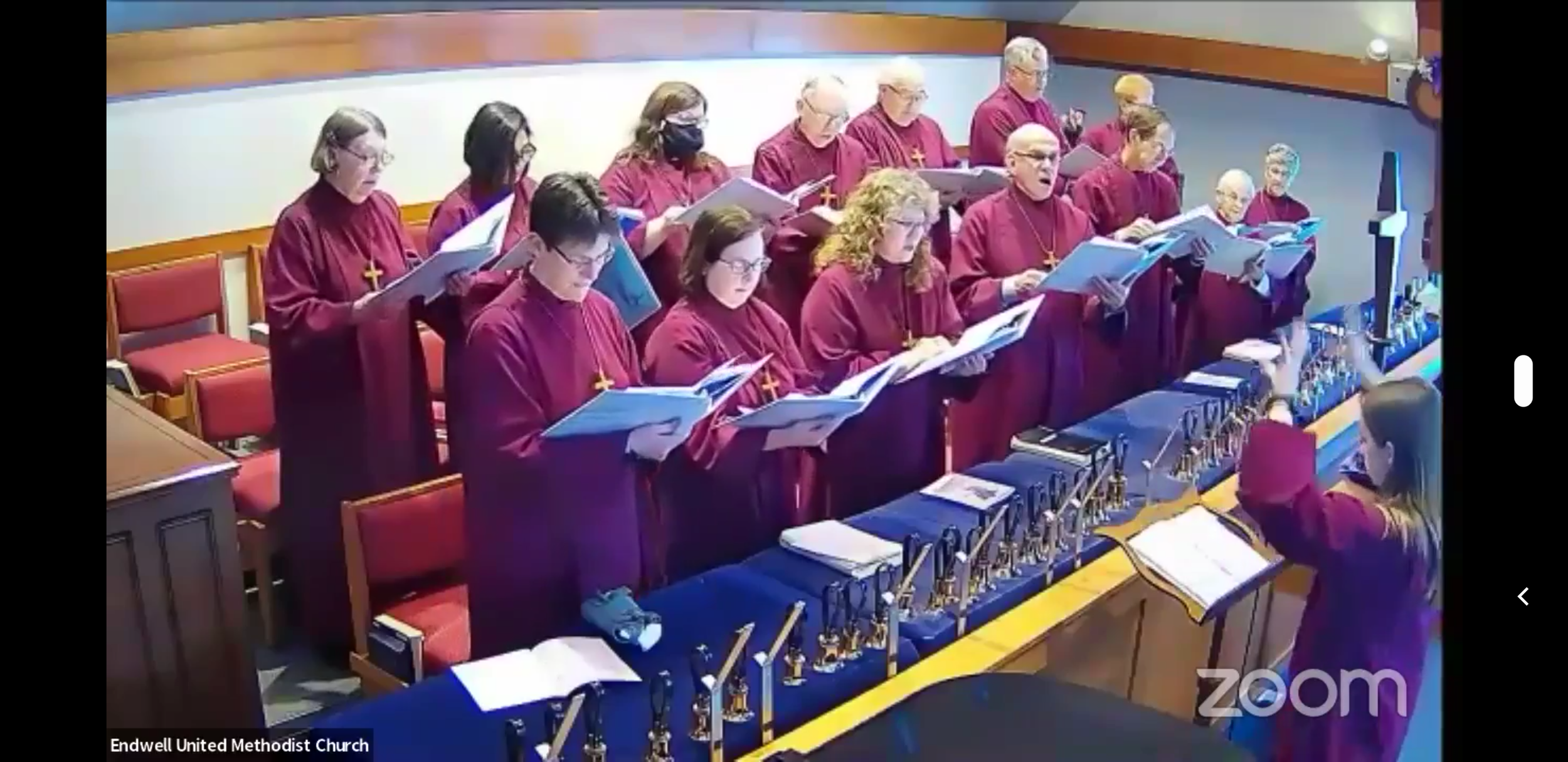A choir of people wearing matching maroon robes with gold crosses singing in a church. They are standing behind a row of bells and music stands, with some holding open songbooks. The church has red cushioned chairs and a wooden railing, and a wooden curved wall in the background.