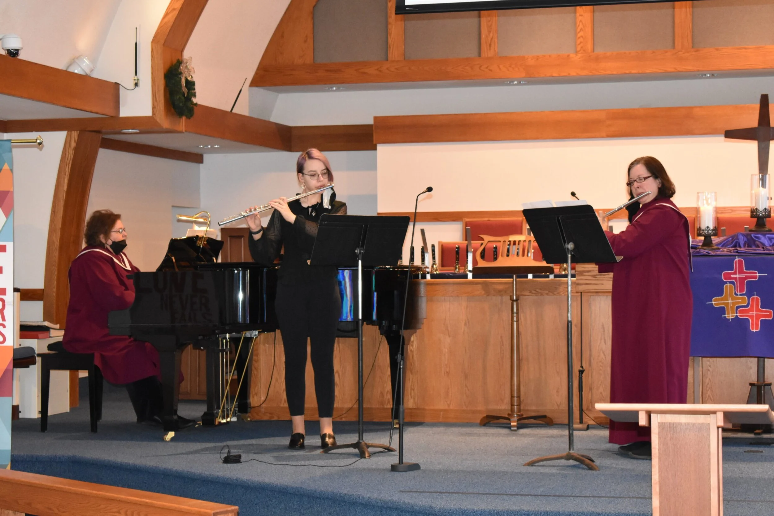 Three women playing musical instruments on a church stage: one on piano, one playing a flute, and one singing or speaking into a microphone. The setting has wooden beams and religious decorations.